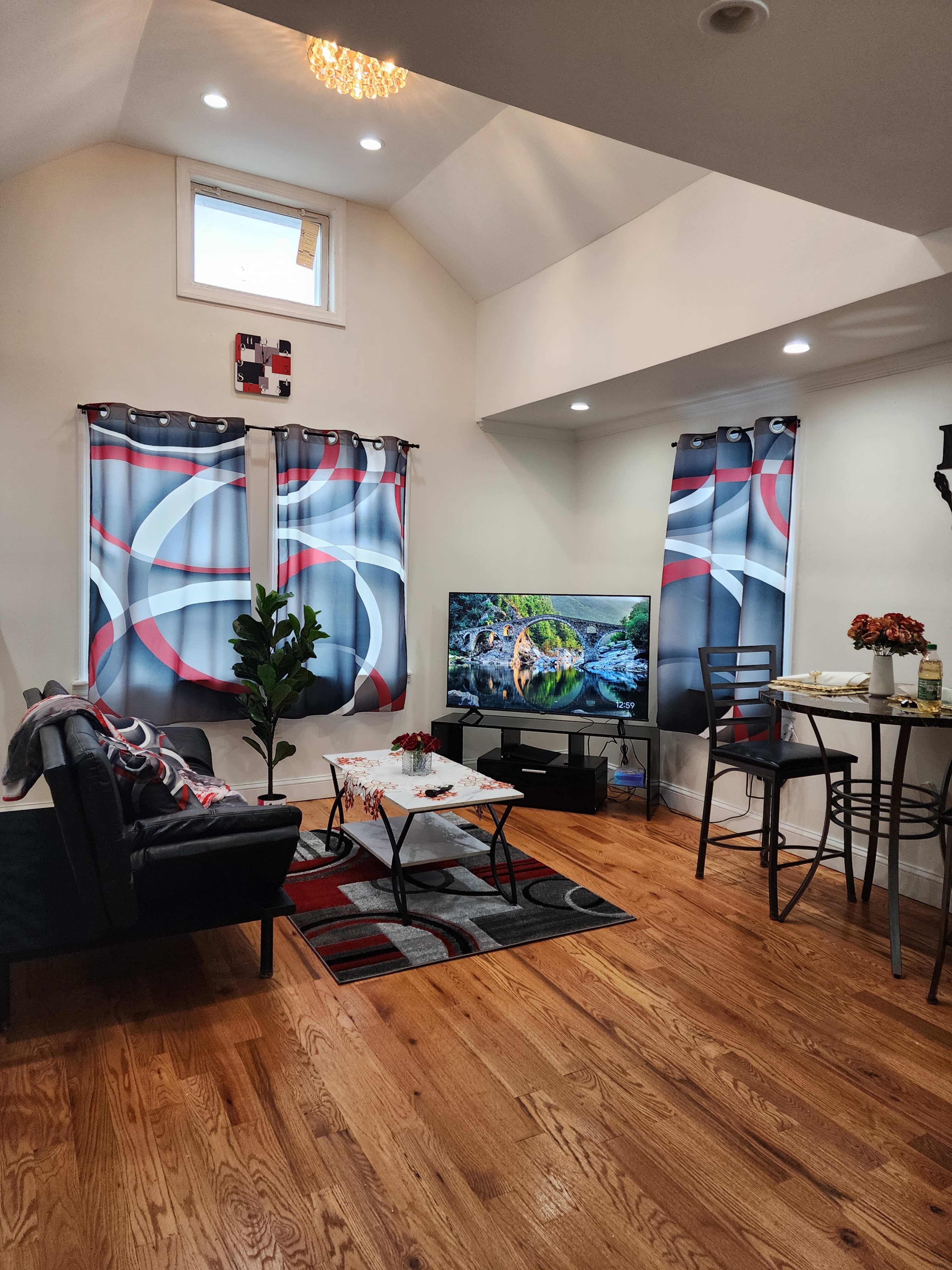 The image shows a cozy living room with a couch, a television displaying a nature scene, and a small dining table with chairs, all under a high ceiling.