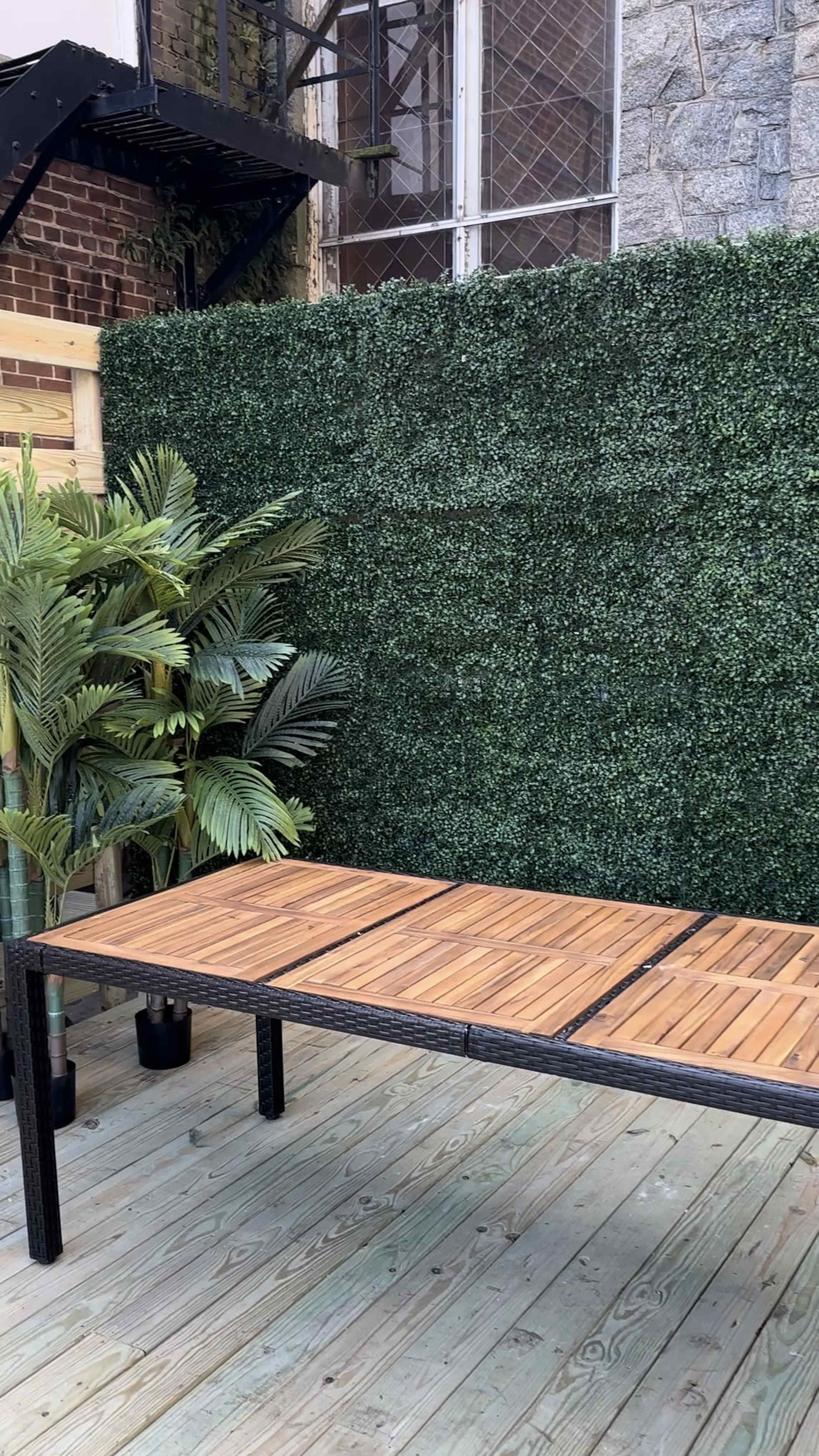 The image features a wooden table in front of a green artificial hedge and potted plants on a wooden deck.