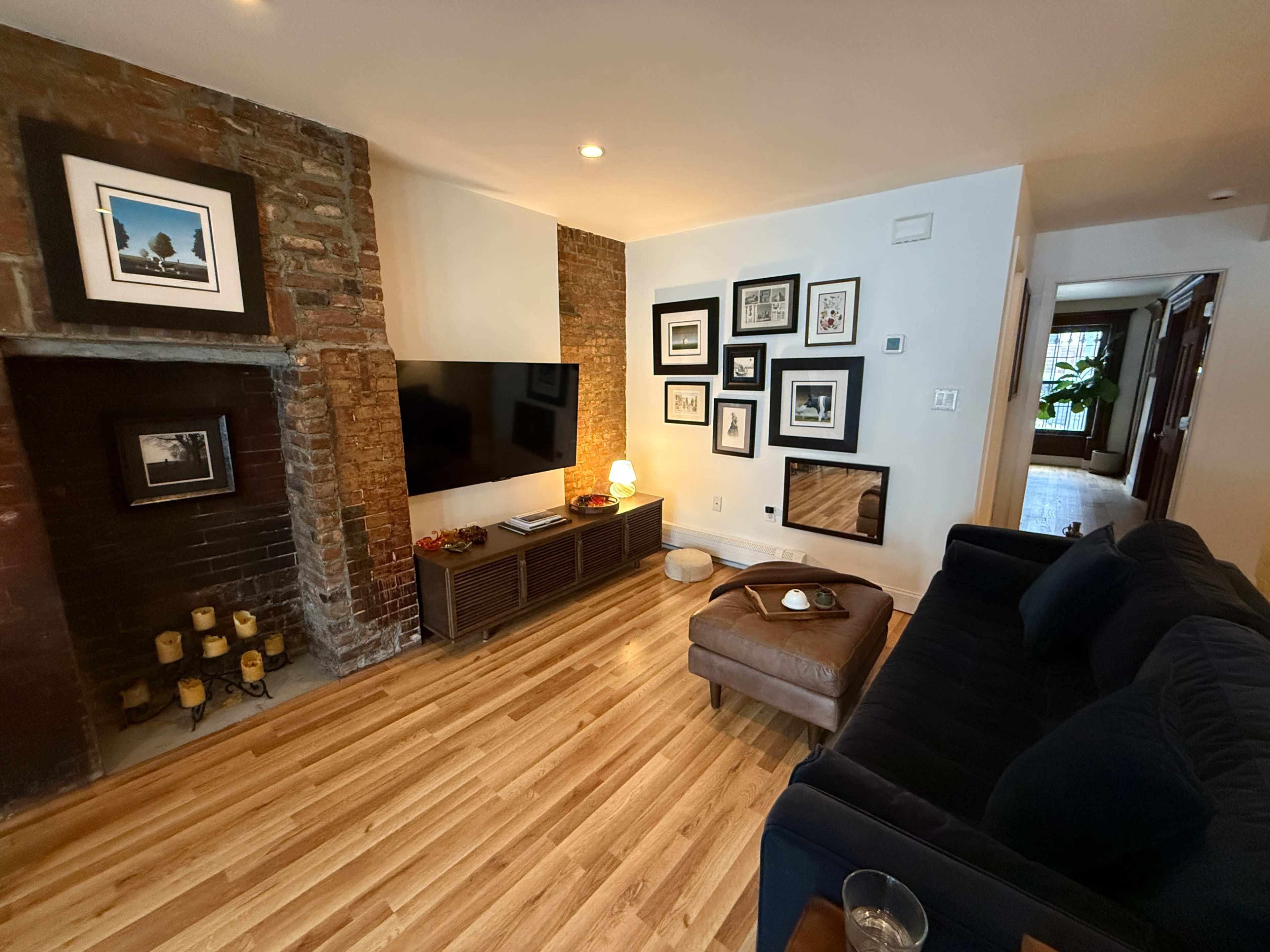The image shows a cozy living room with a brick feature wall, a mounted television, framed photos on the wall, a leather ottoman, and a dark blue sofa.
