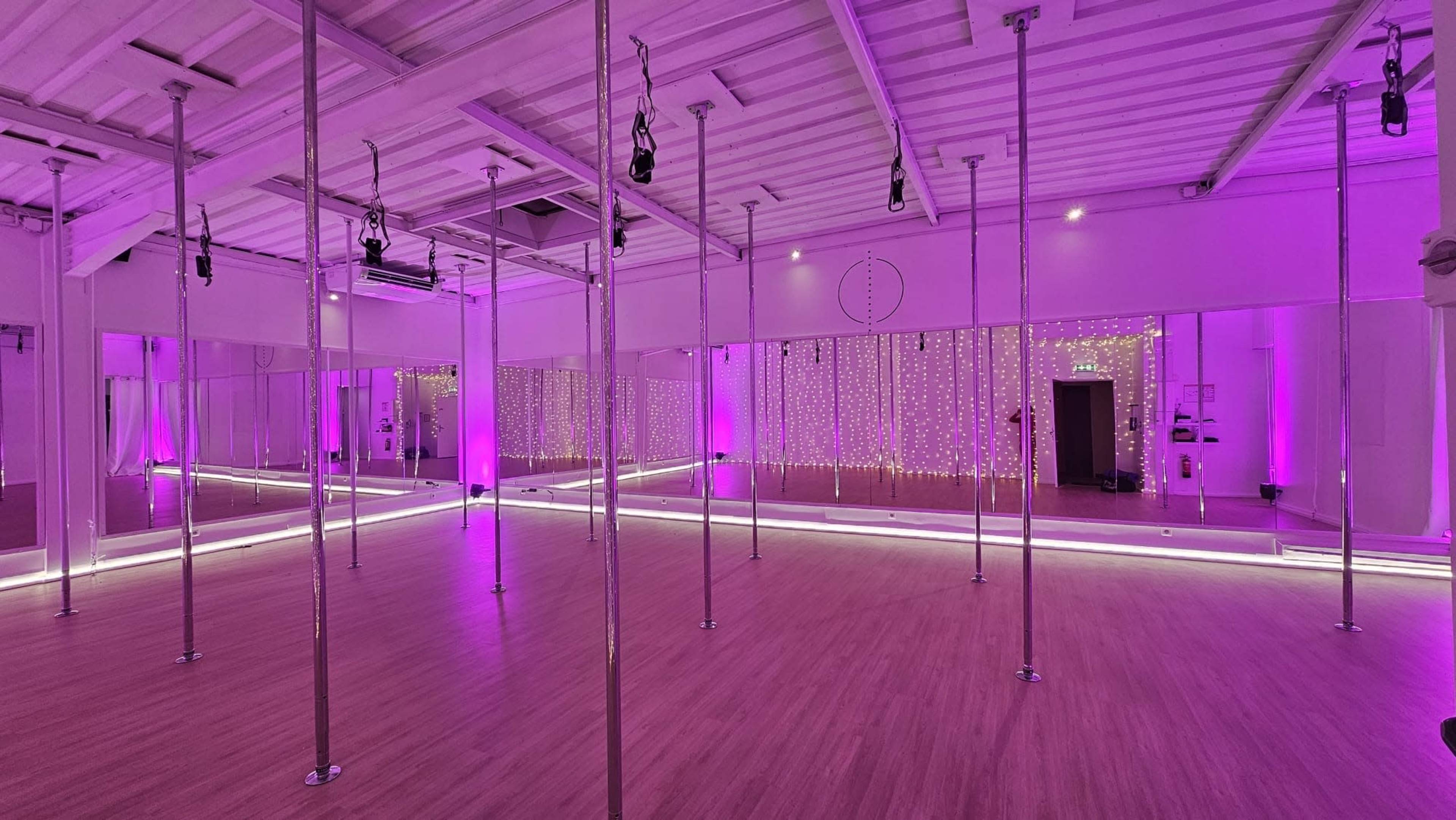 A spacious dance studio features polished wooden floors, multiple poles, and mirrors, illuminated by purple lights and decorative strings of lights.