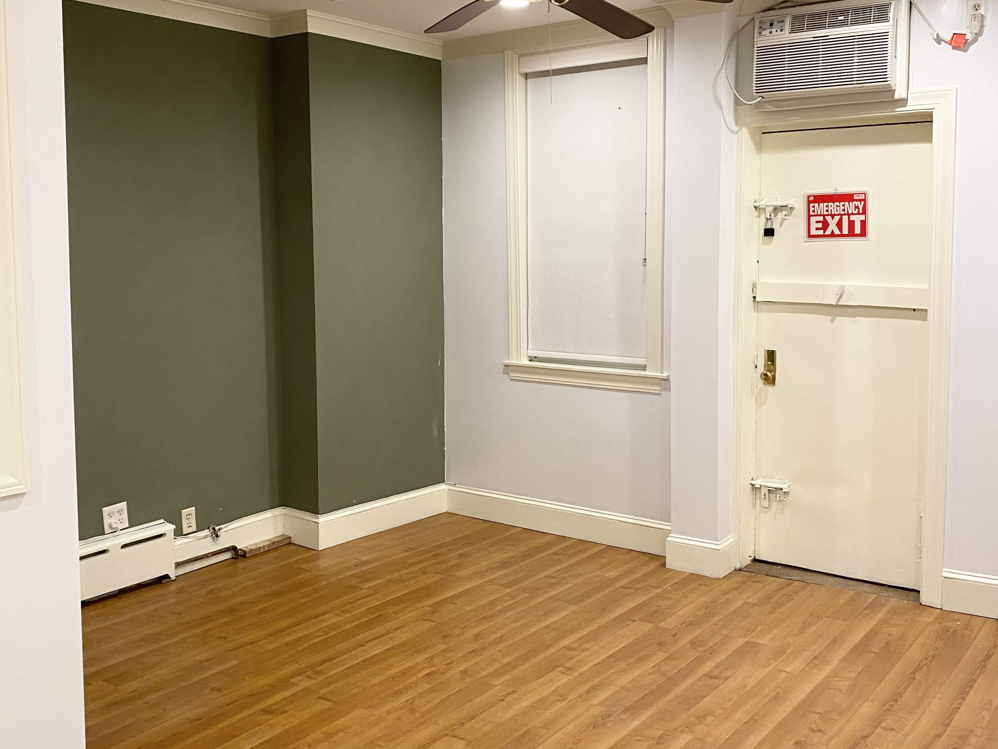 The image shows a small room with a green accent wall, a wooden floor, and a door marked "EMERGENCY EXIT."