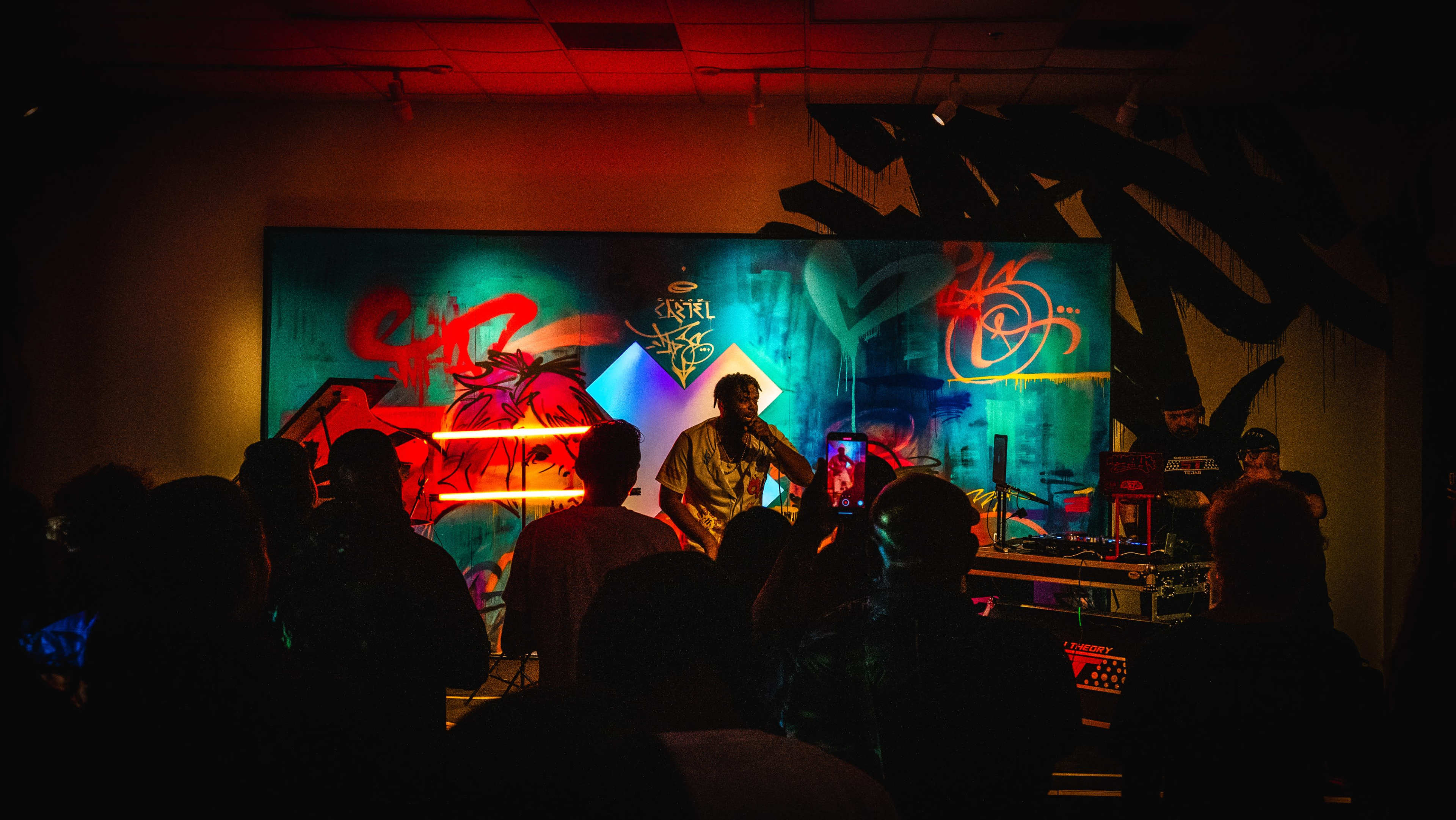 A musician performs on stage in front of a crowd, with vibrant graffiti artwork displayed behind them.