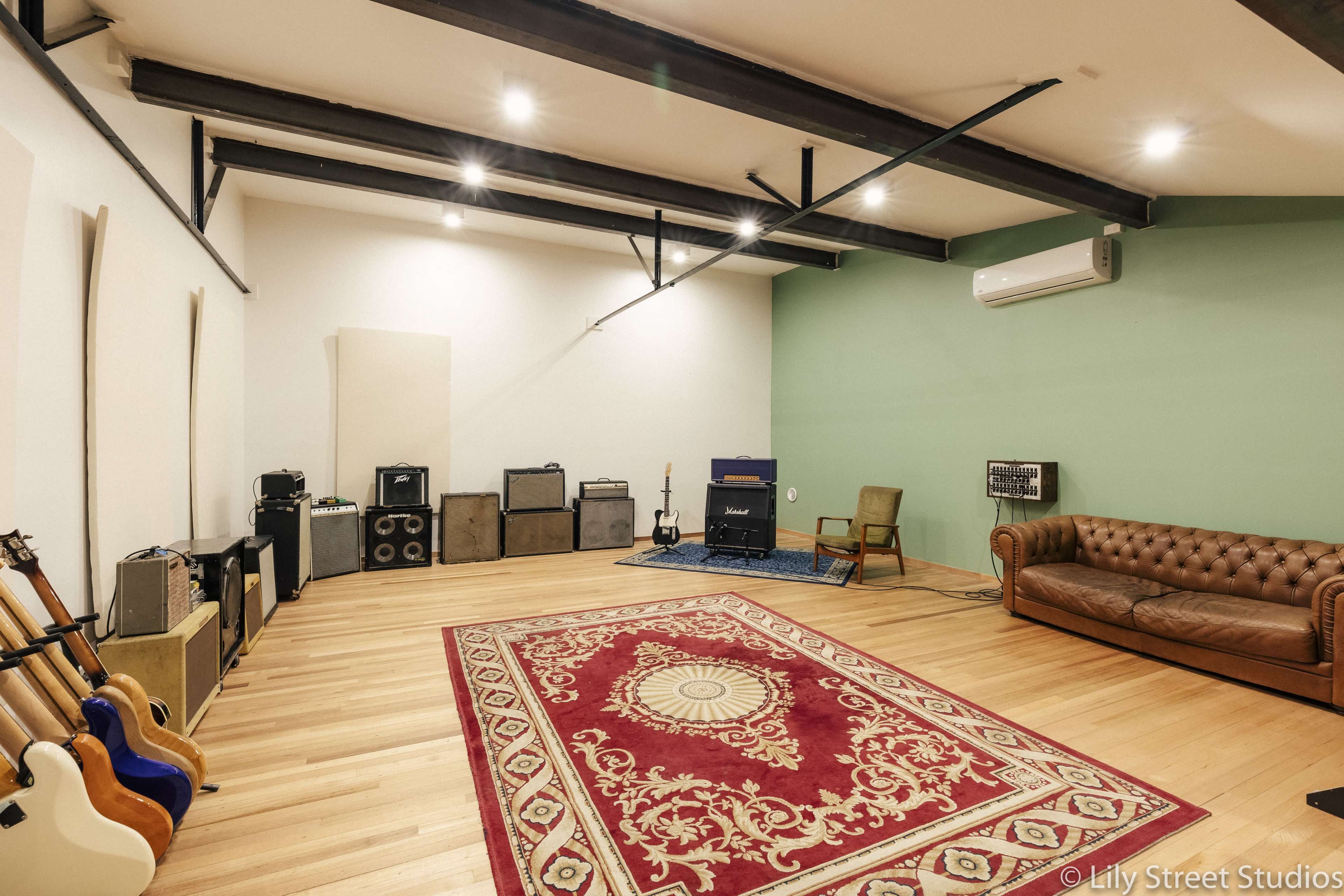 A spacious music rehearsal room with wooden floors, a large rug, an array of amplifiers, and a brown leather couch.