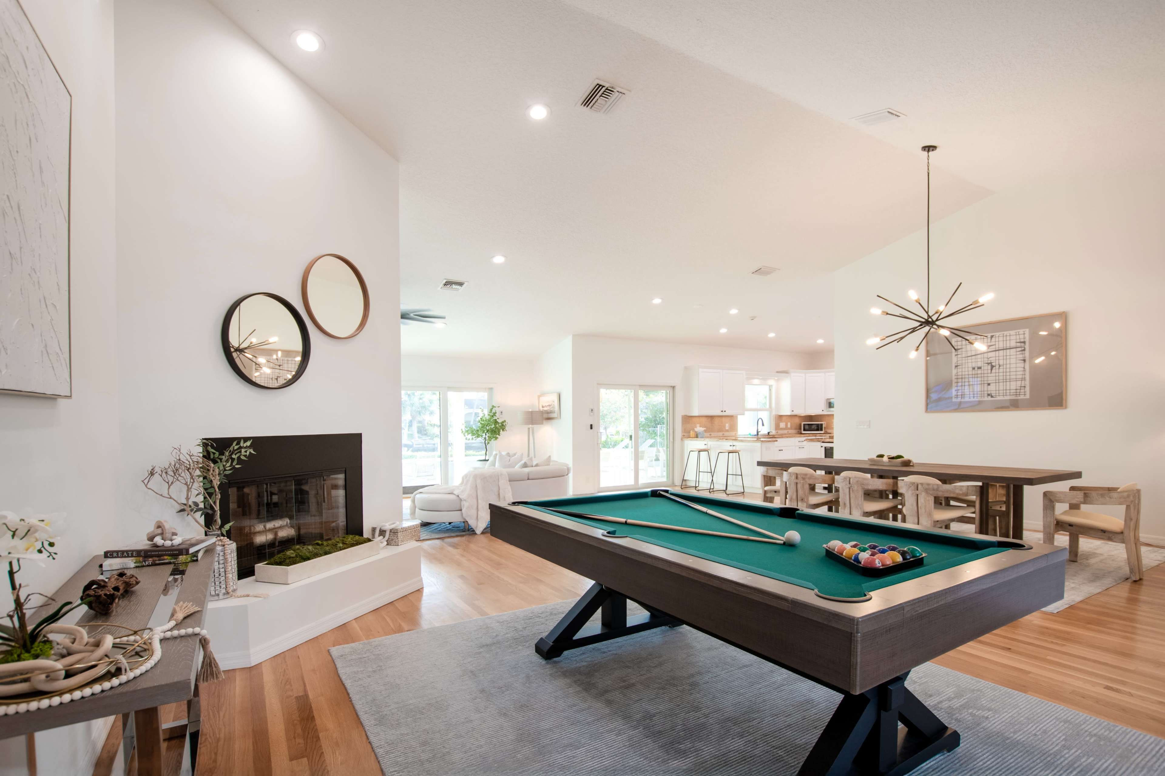 The image shows a modern living space that features a billiard table, a dining area with wooden chairs, a fireplace, and large windows that allow natural light to enter.