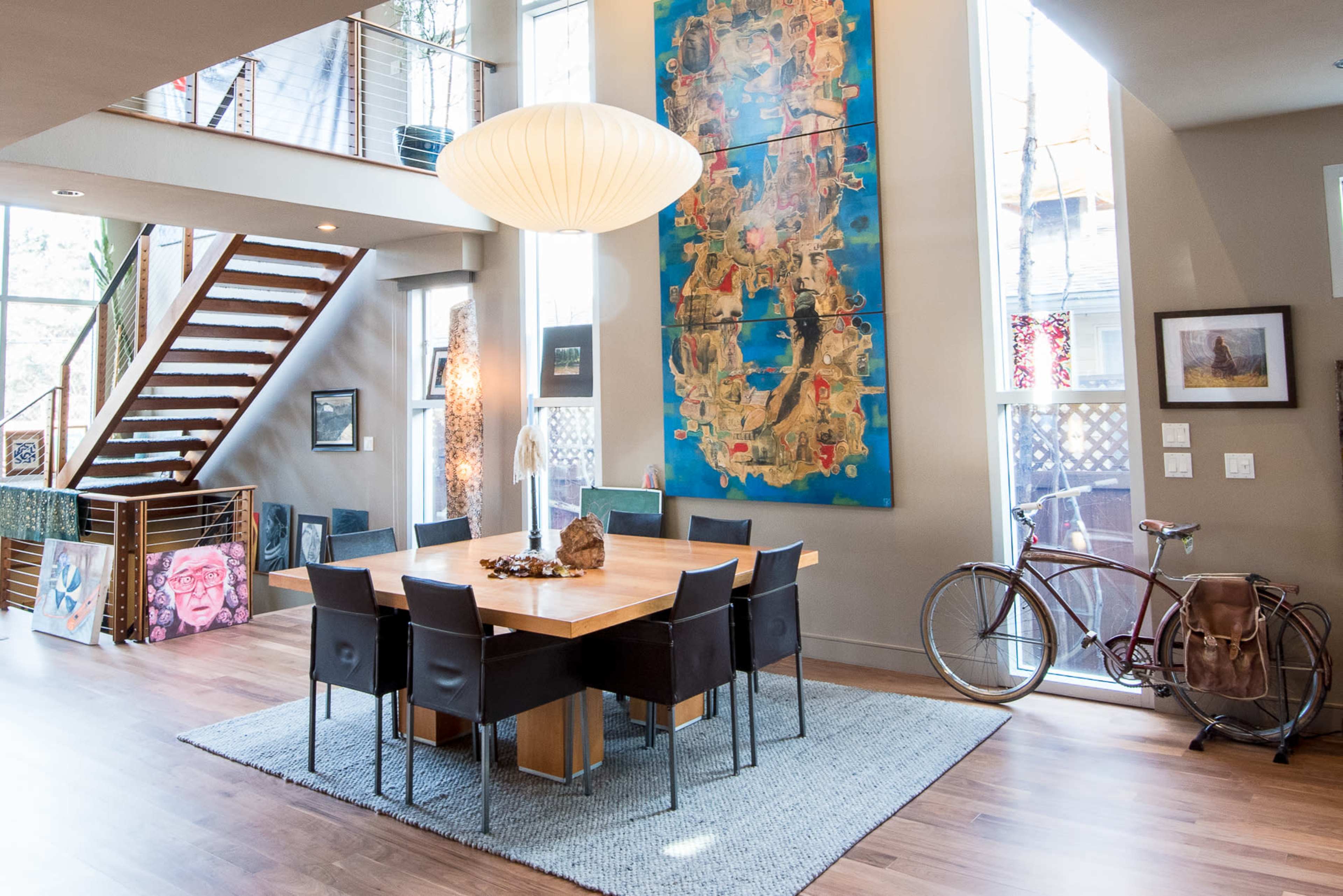 The image shows a modern dining area with a large wooden table surrounded by eight black chairs, a colorful piece of artwork on the wall, and a staircase leading to an upper level.
