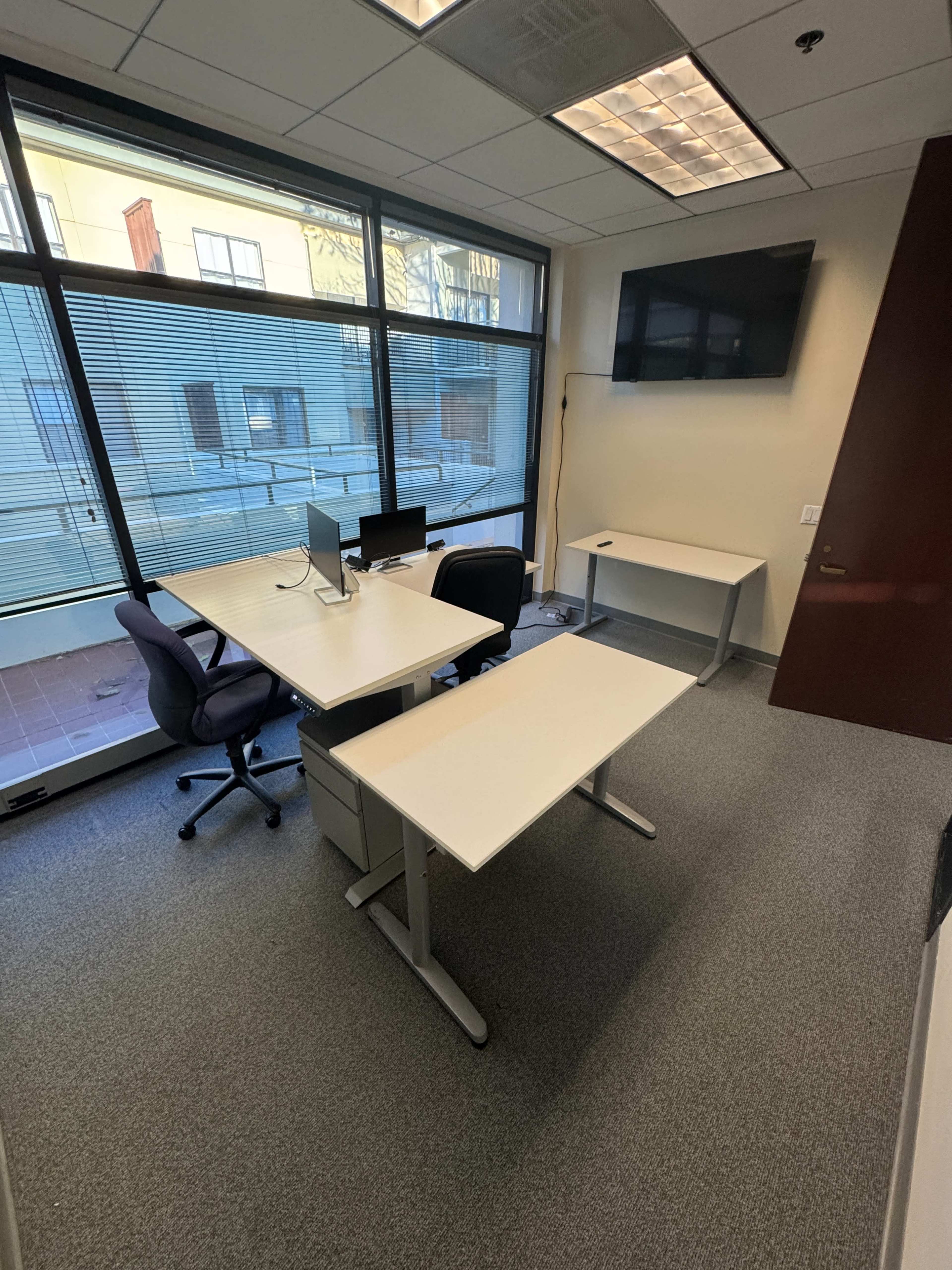 The image shows a small office space with a desk, chair, computer setup, and a television mounted on the wall, all positioned near large windows.