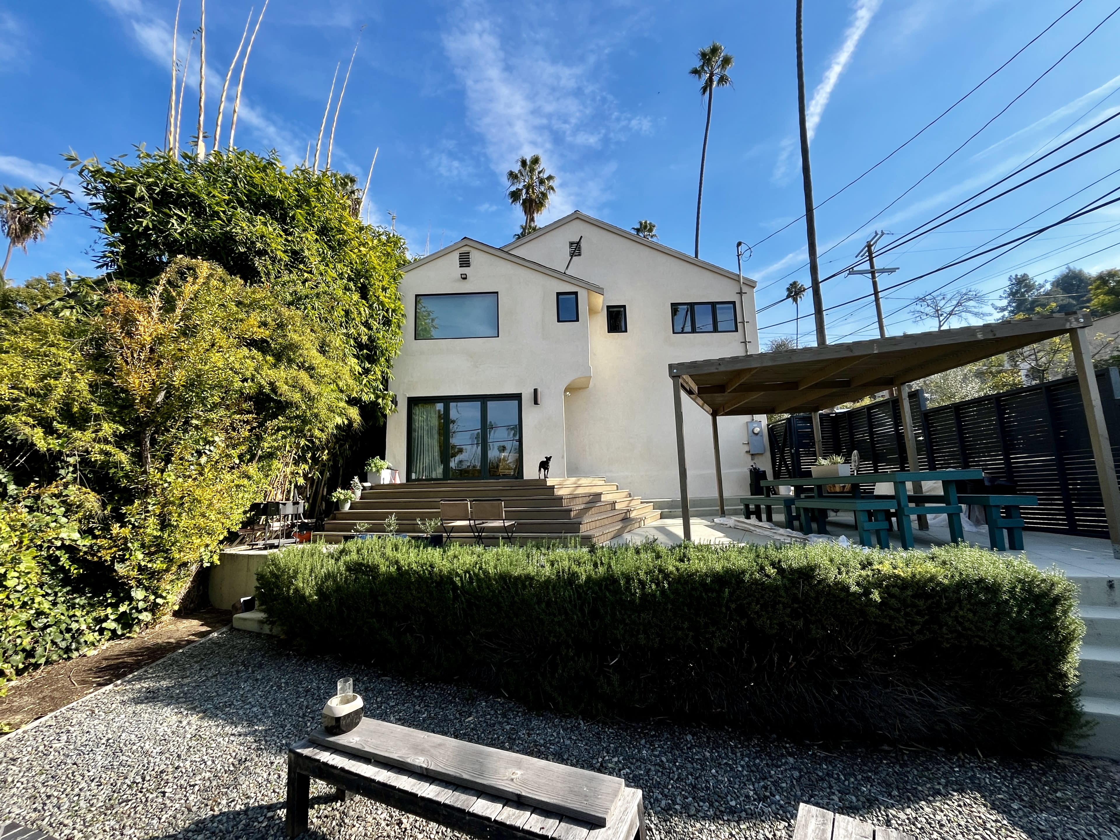 The image shows a modern house with a landscaped garden, featuring wooden steps and a patio area, surrounded by palm trees and power lines.