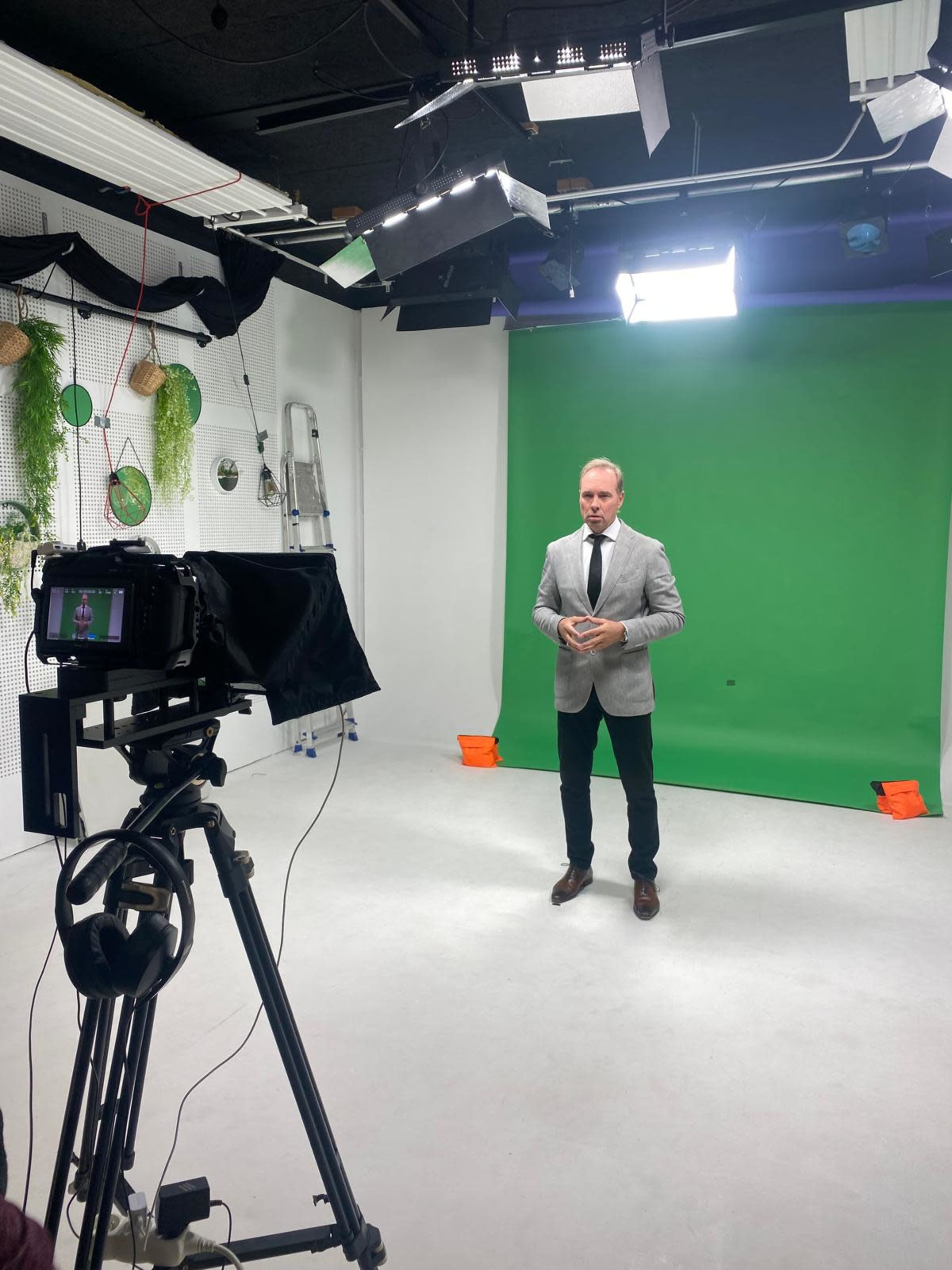 A man in a gray suit stands on a white studio floor in front of a green backdrop, with a camera and lighting setup nearby.