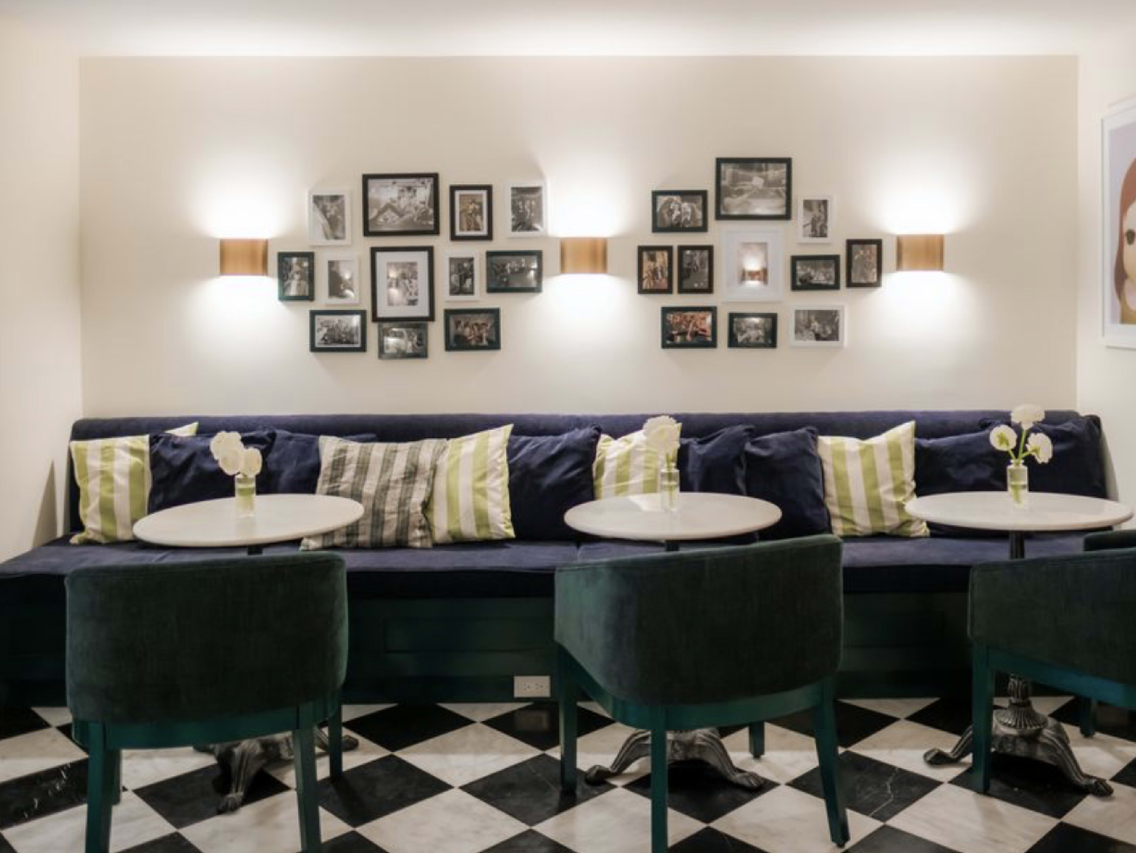 The image shows a cozy café seating area with a dark blue upholstered bench, round tables, and green velvet chairs, accompanied by black and white photographs on the wall.