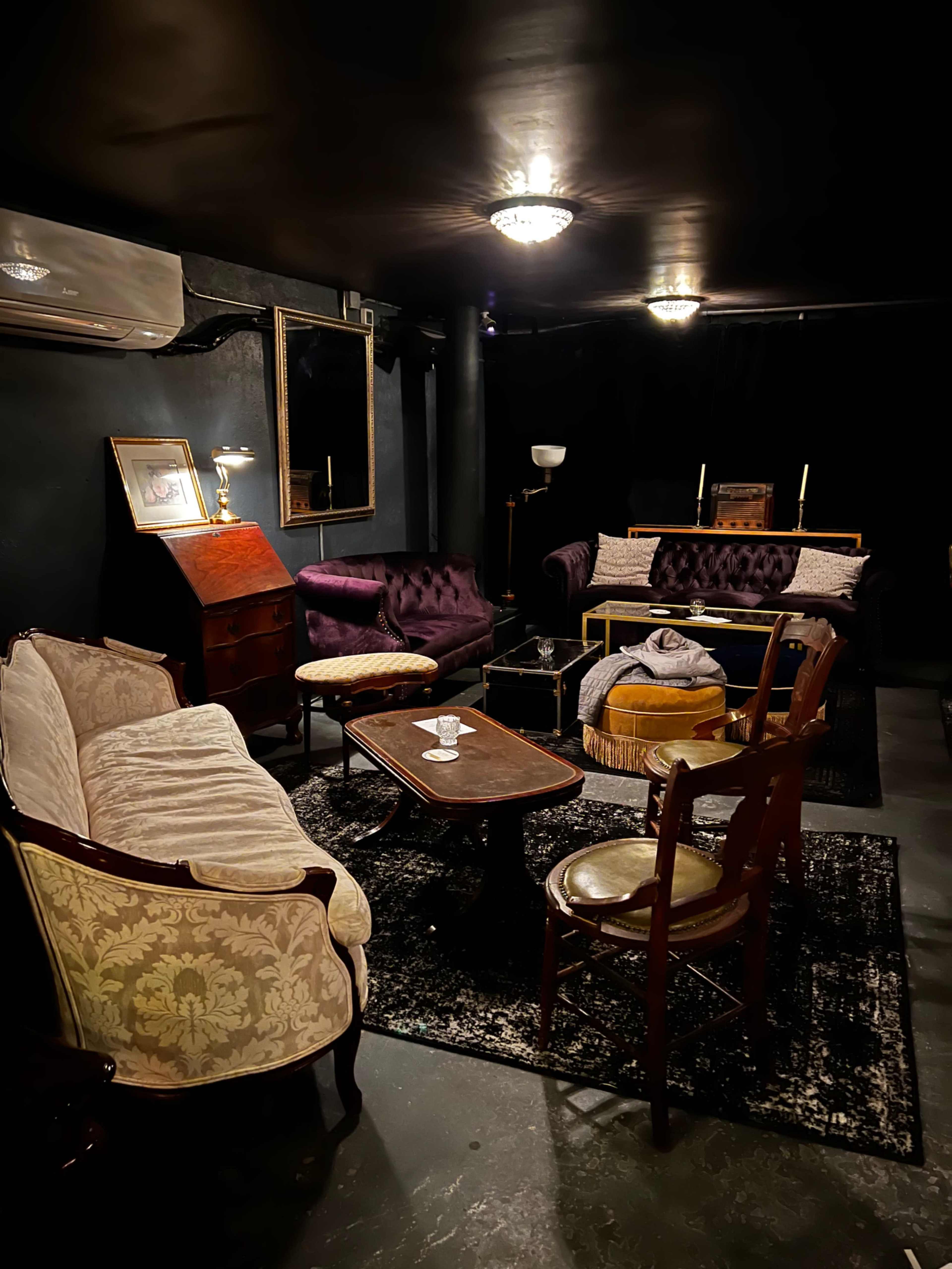A dimly lit lounge area with various vintage-style seating, a small table, and a dark rug on a concrete floor.