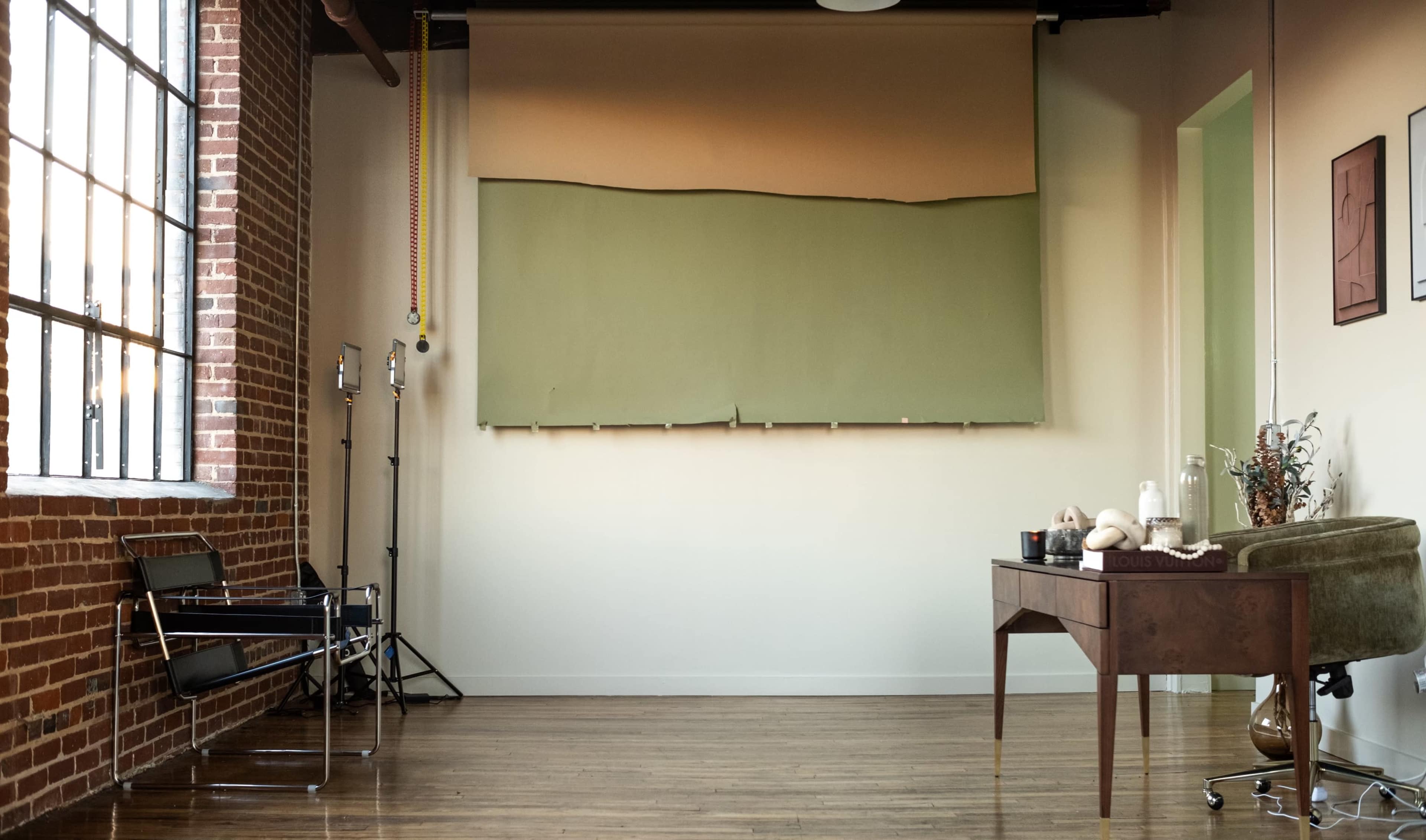 A spacious room features a large window, a green backdrop mounted on the wall, and a wooden desk with decorative items.