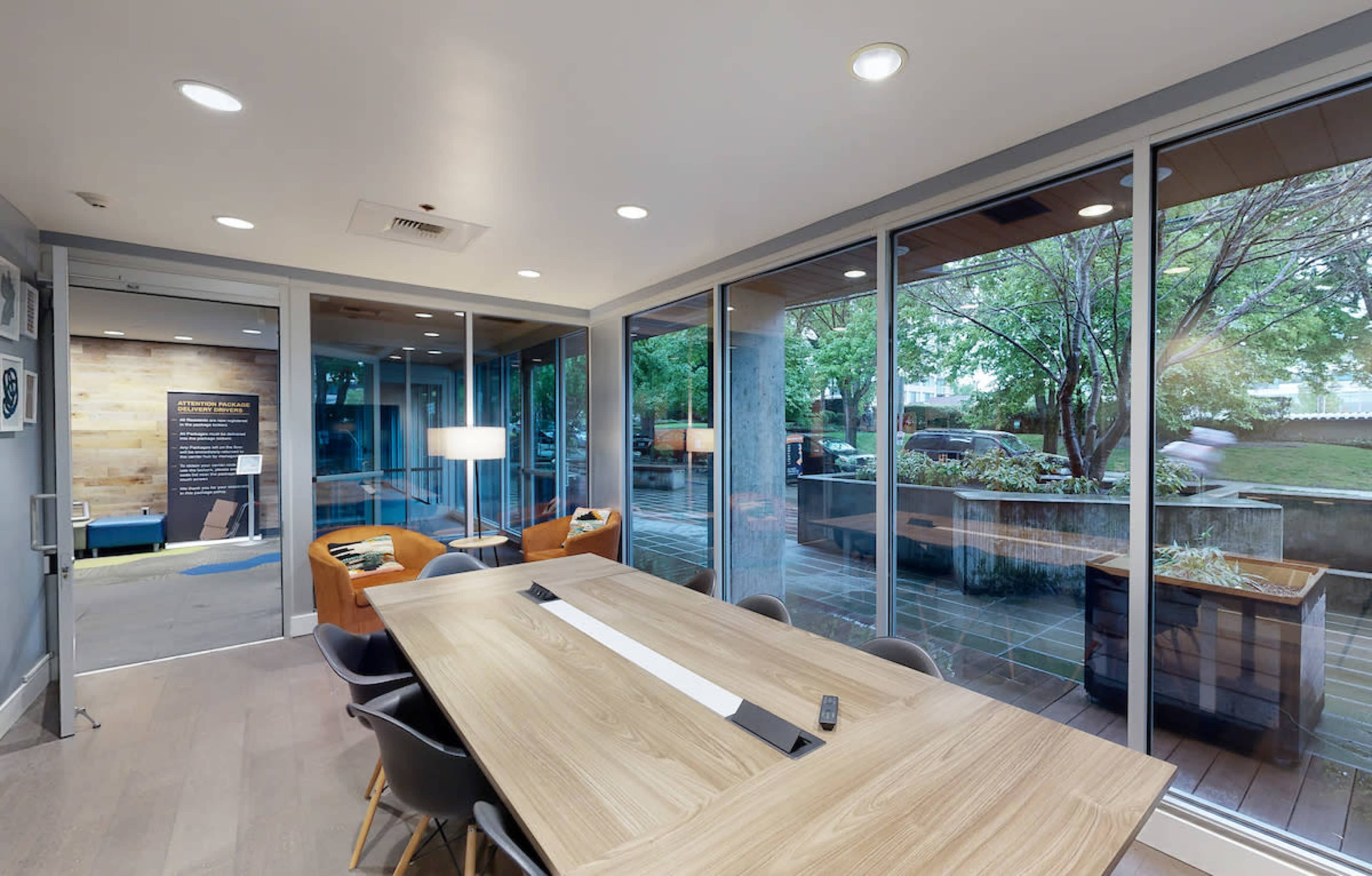 A modern conference room with a large wooden table, surrounded by chairs, and large glass windows that overlook a landscaped outdoor area.