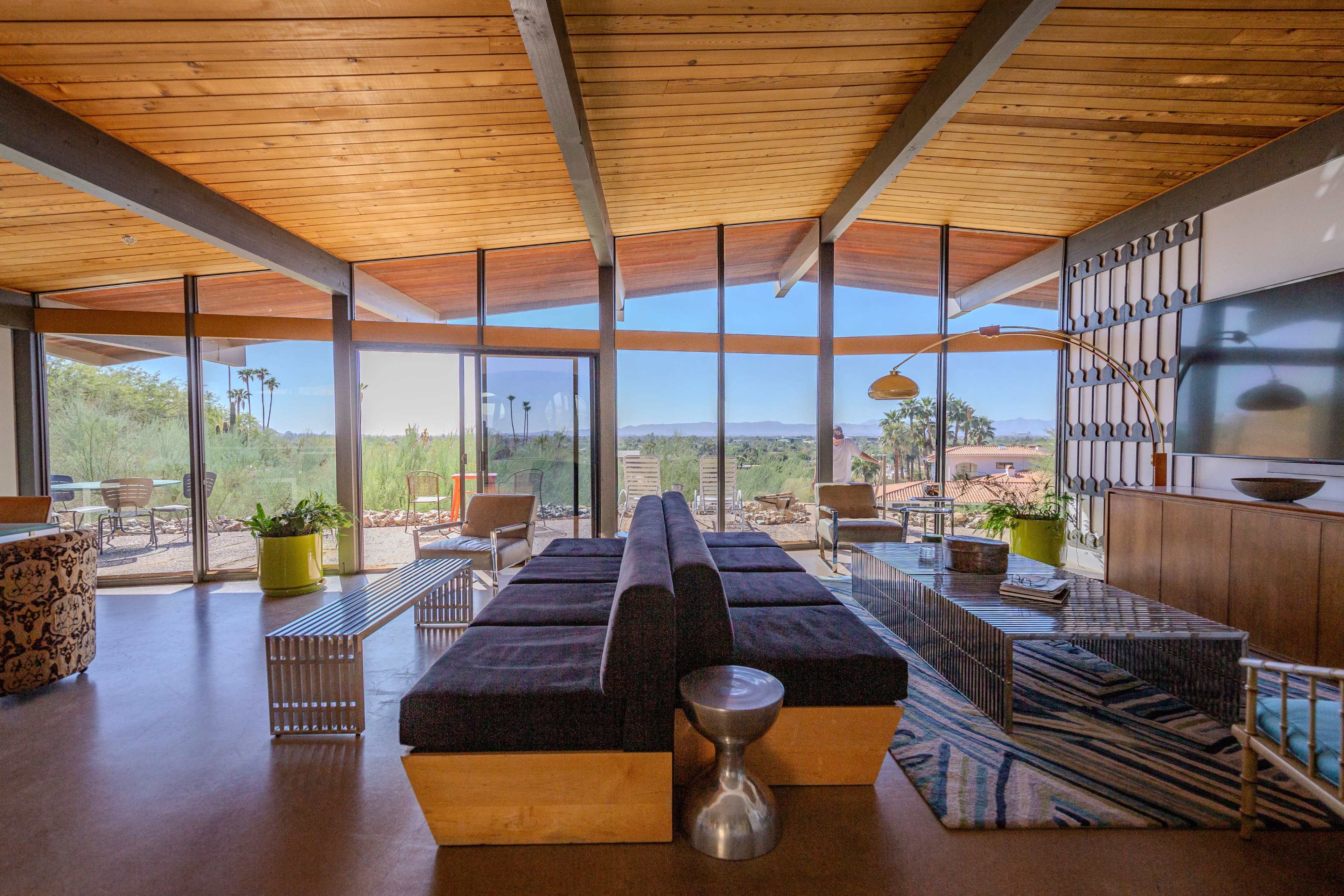 The interior of a modern living space features a large sofa, a metal side table, and expansive windows that showcase a desert landscape.