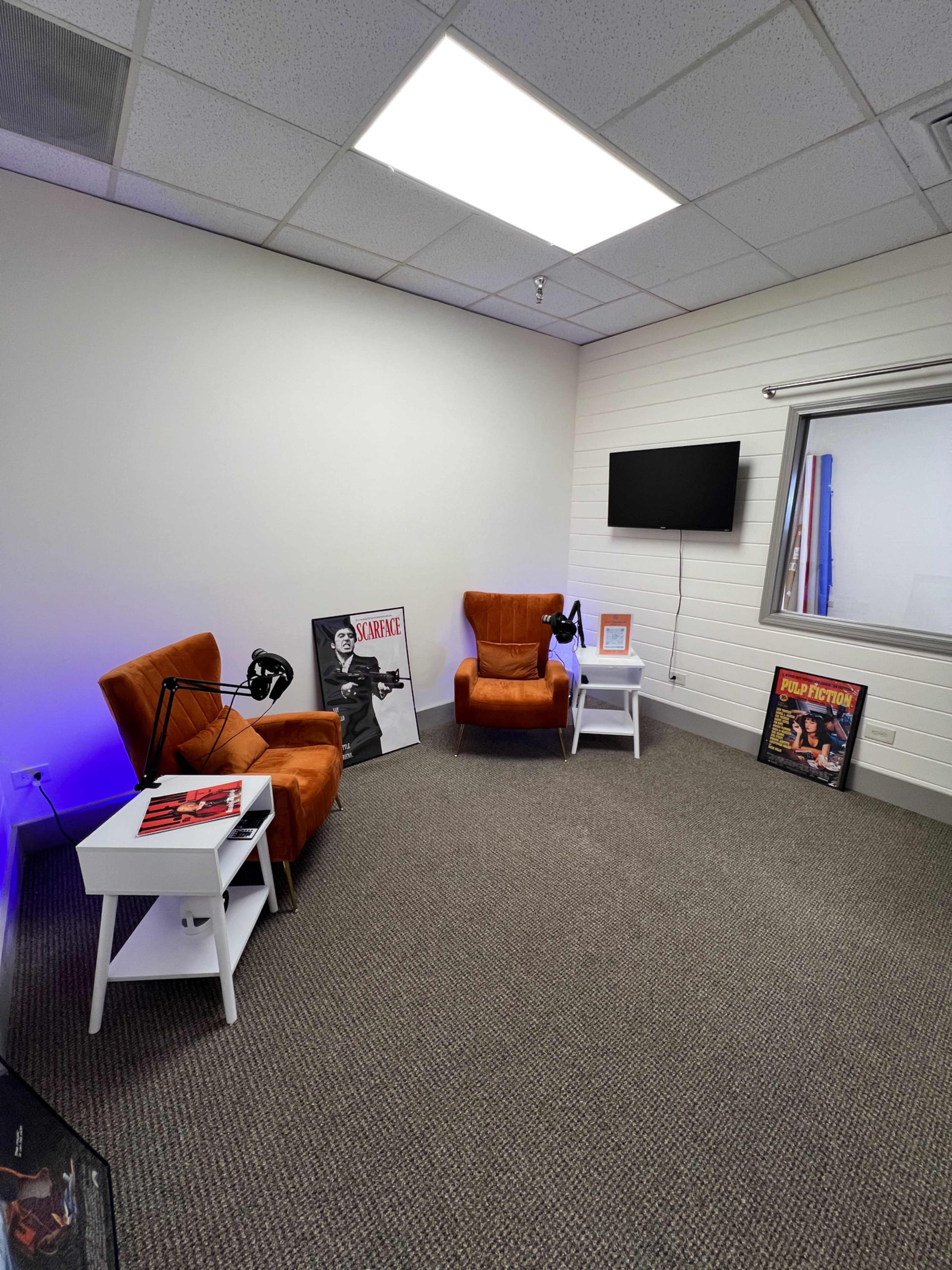 The image shows a minimalistic room with two orange armchairs, a small white side table, a wall-mounted TV, and posters in frames on the floor.