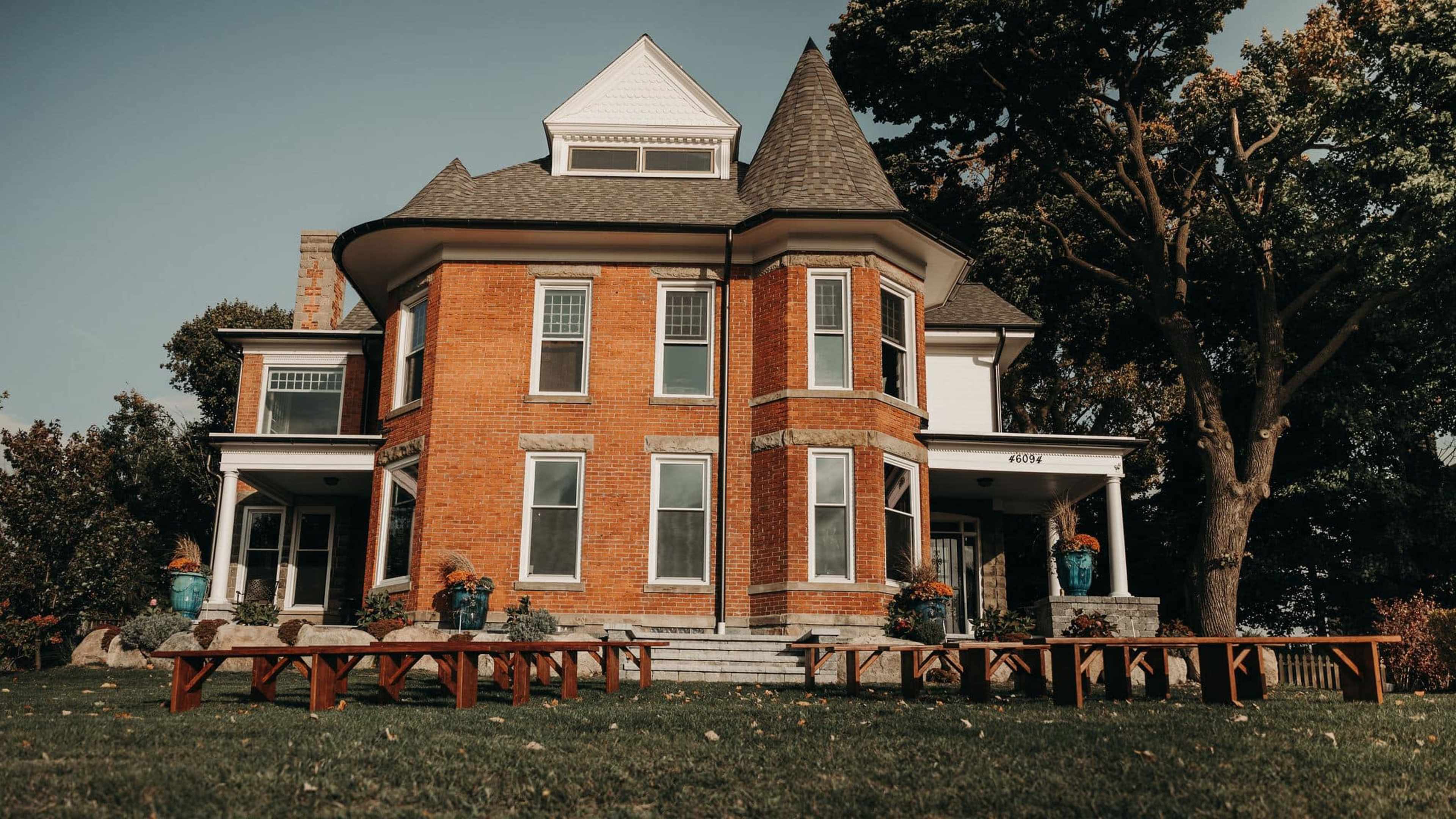 A Victorian-style brick house features a conical roof and large windows, surrounded by grass and wooden benches.