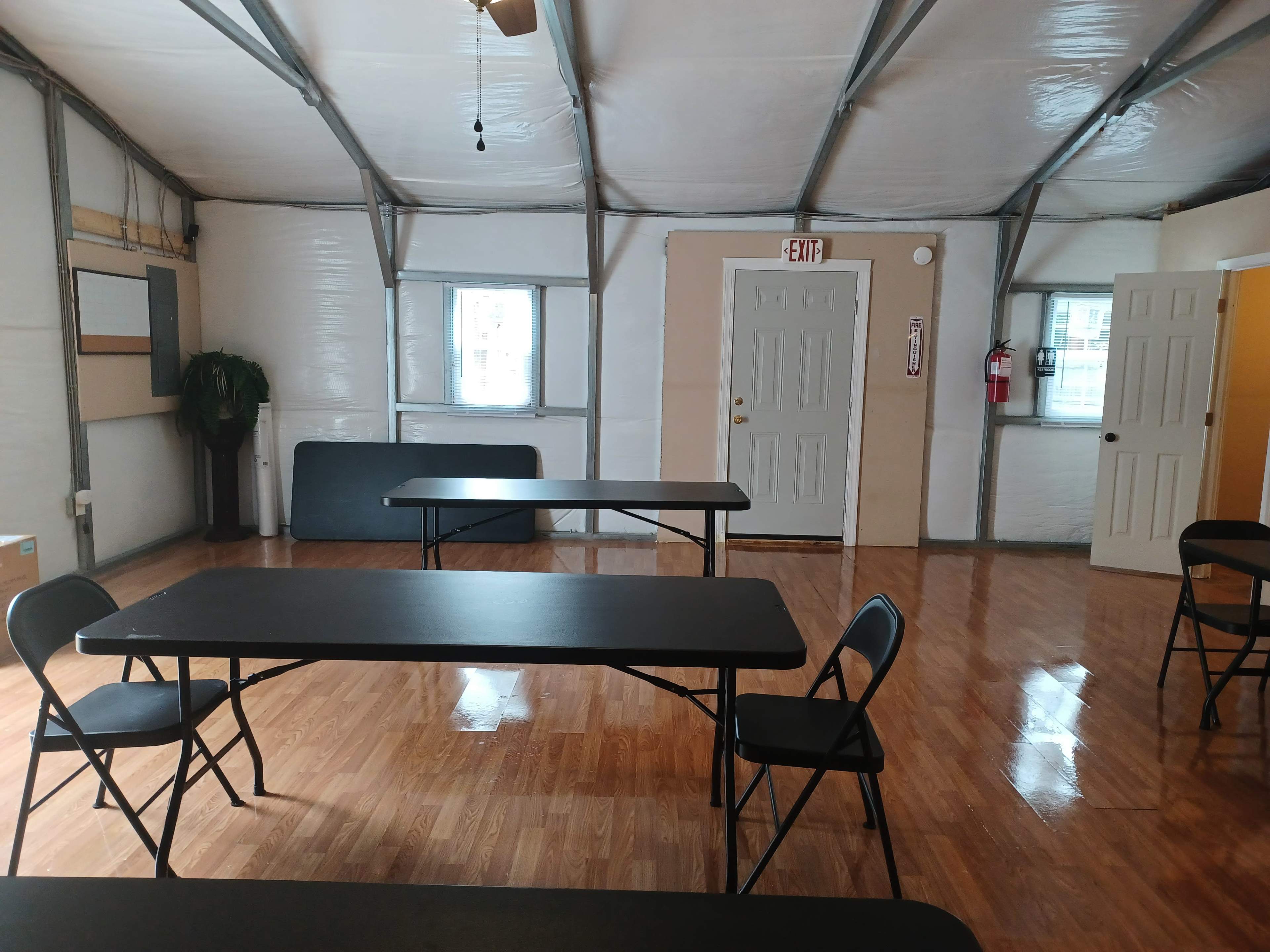 The image shows a spacious indoor area with several black folding tables and chairs, and two doors along the walls.