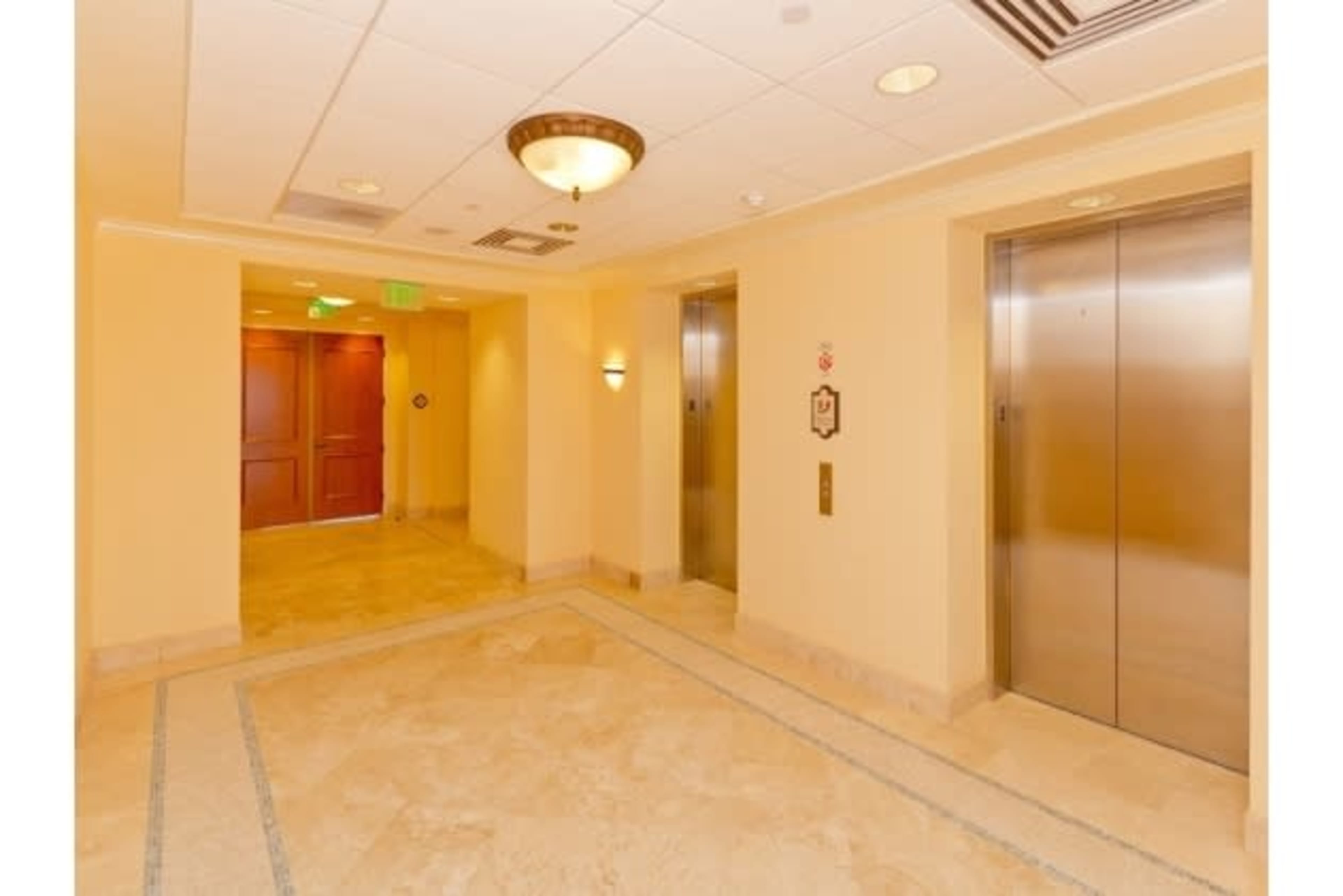 A hallway with two elevator doors on the right and a wooden door leading to another room on the left, all set on a light-colored tiled floor.