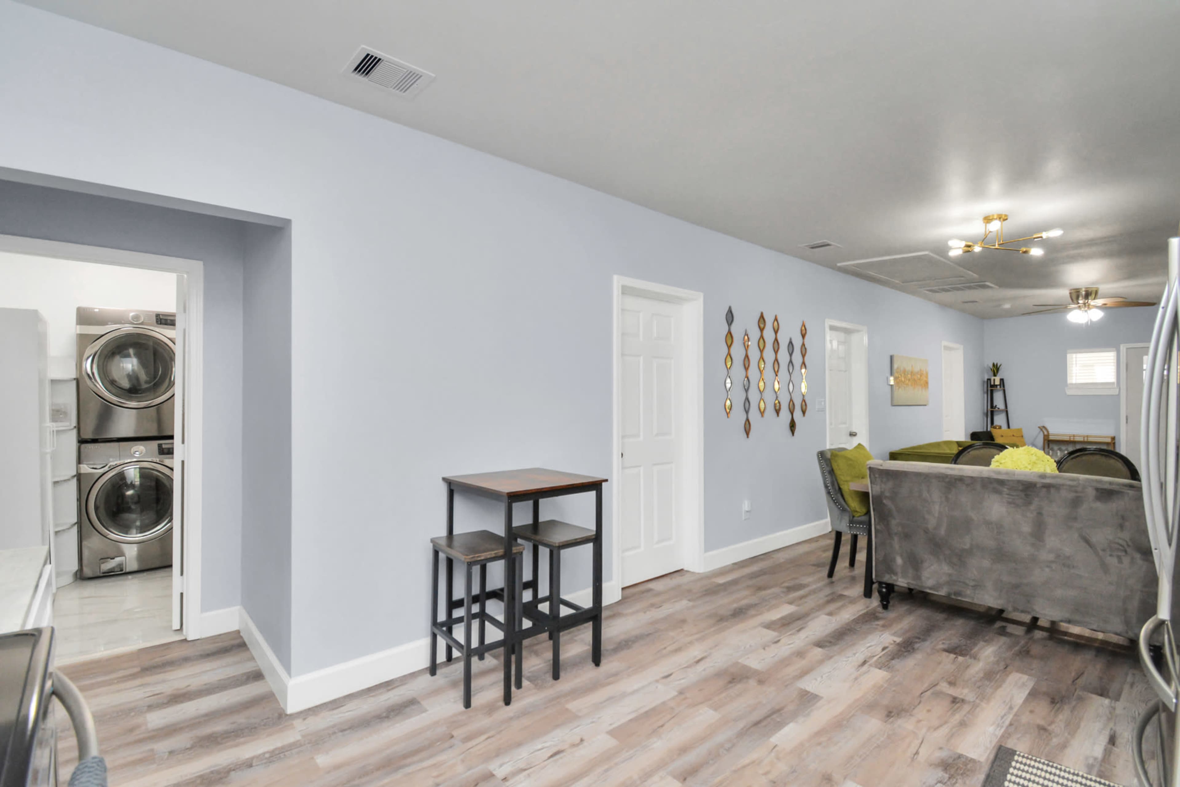 A light blue interior space featuring a small table with two chairs, a living area with gray furniture, and a laundry area visible through an open door.