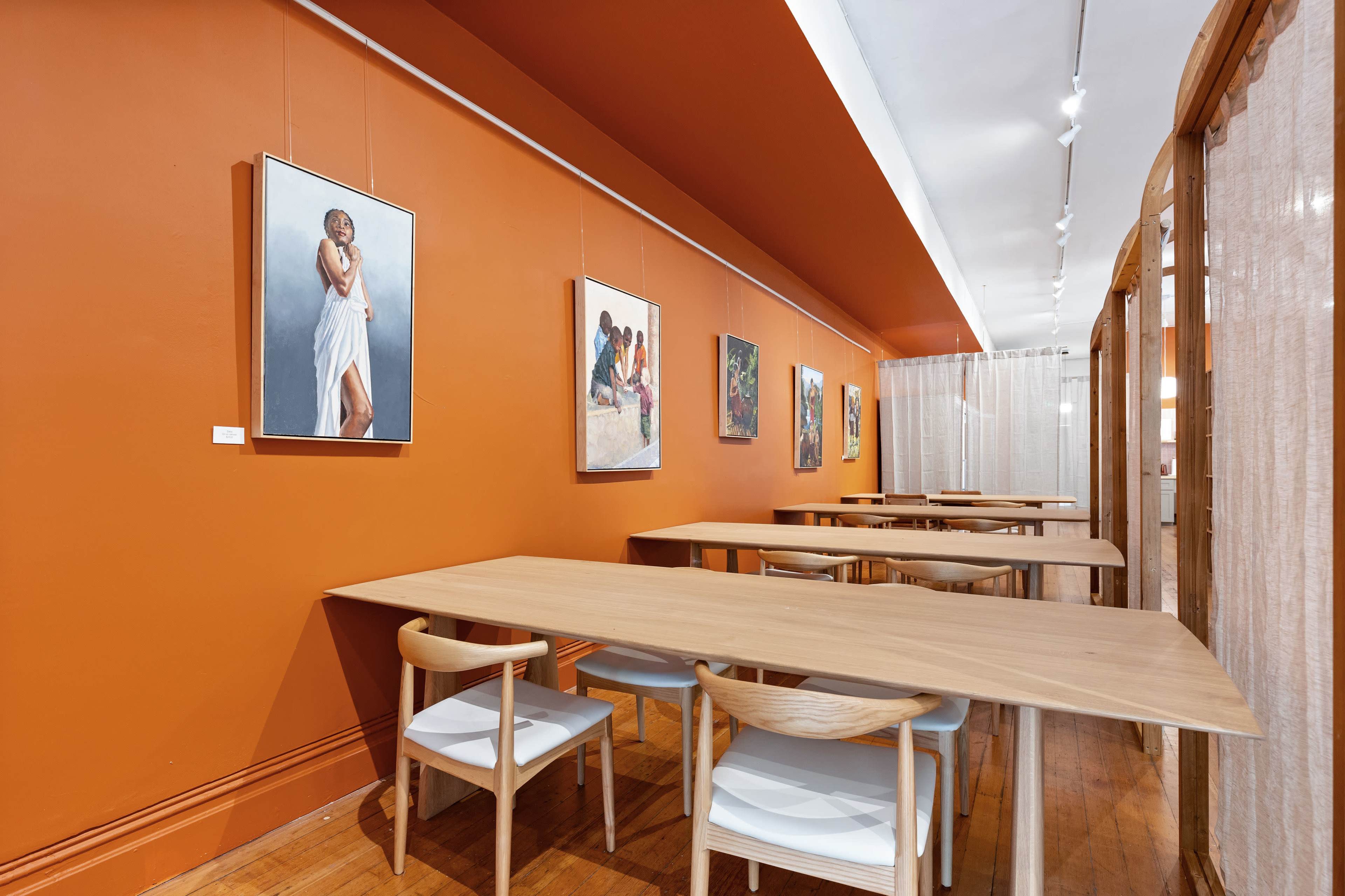 A modern art gallery featuring an orange wall with several paintings and wooden tables arranged in a brightly lit space.