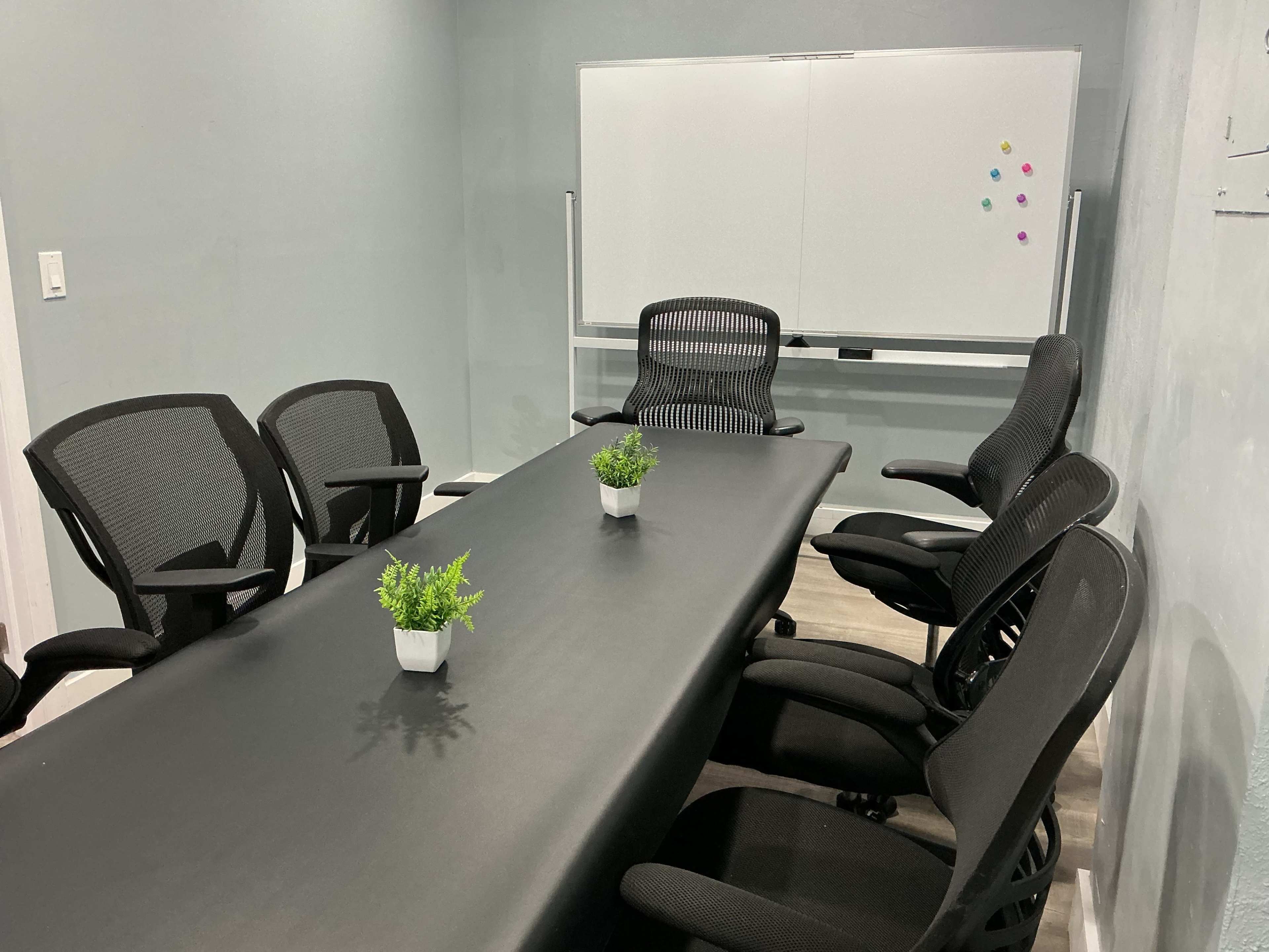 A conference room features a long black table surrounded by six ergonomic chairs, with two small plants on the table and a whiteboard in the background.
