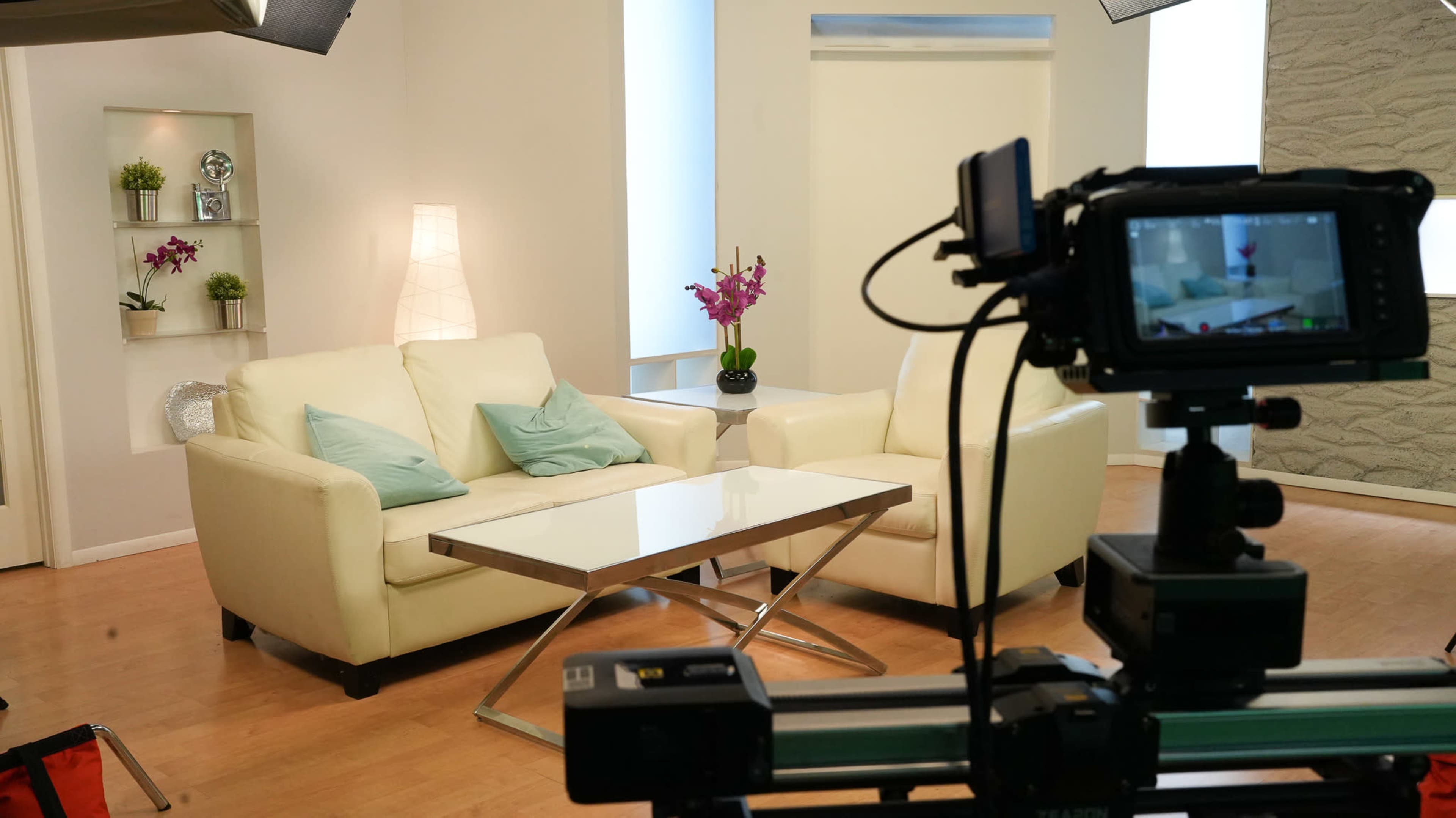 A white sofa with light blue cushions is positioned in a well-lit studio, facing a modern glass coffee table and decorated with potted orchids.