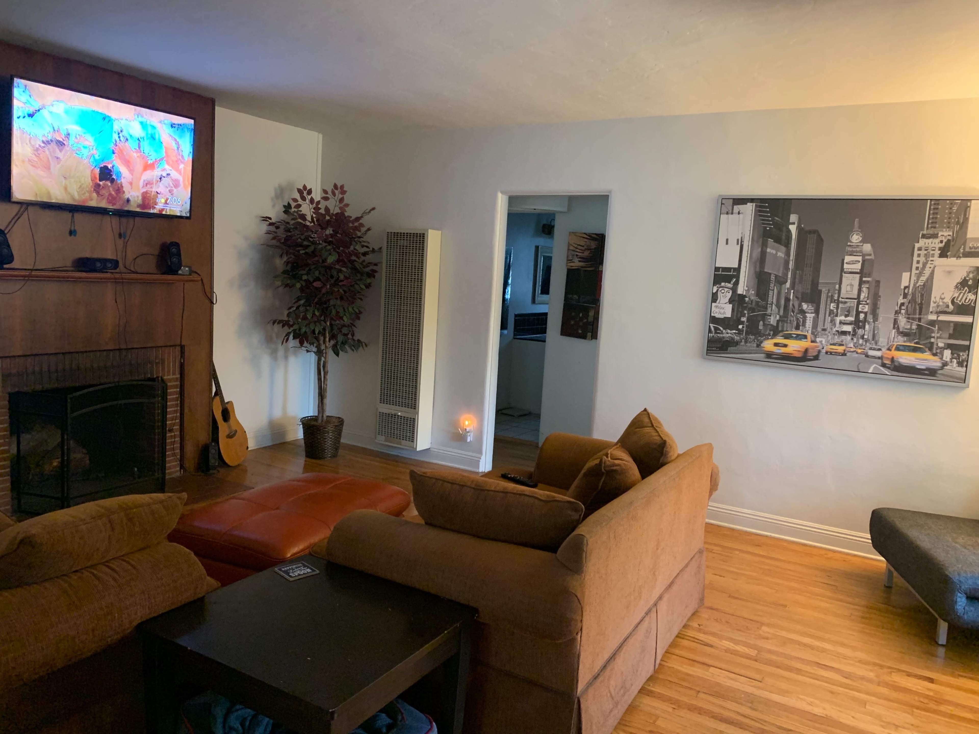 A cozy living room with a brown sectional sofa, a coffee table, a television mounted on the wall, and a large photo of a city street featuring yellow taxis.