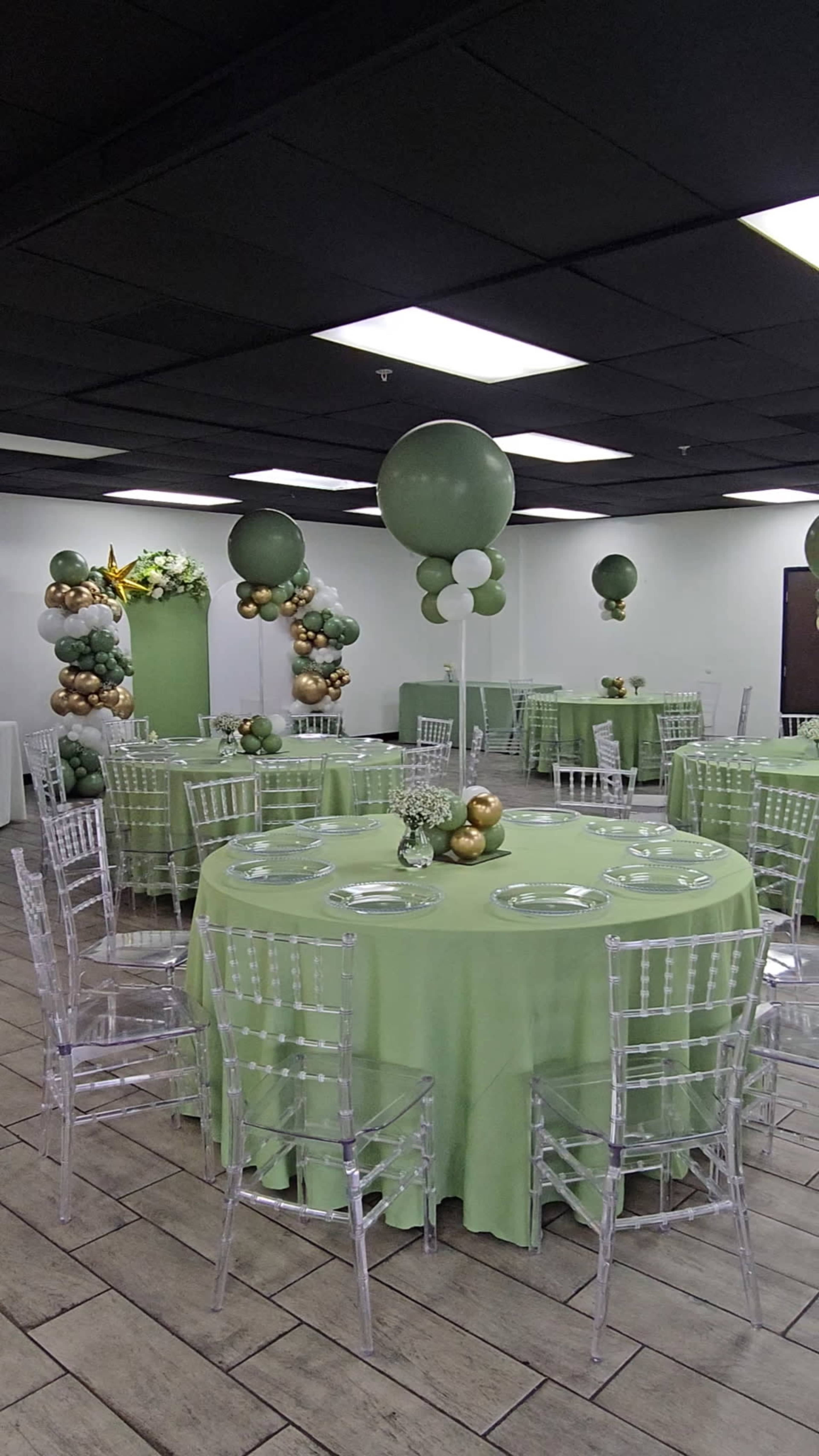 The image shows a decorated interior space with several round tables covered in light green tablecloths, transparent chairs, and a balloon arch in shades of green and gold.