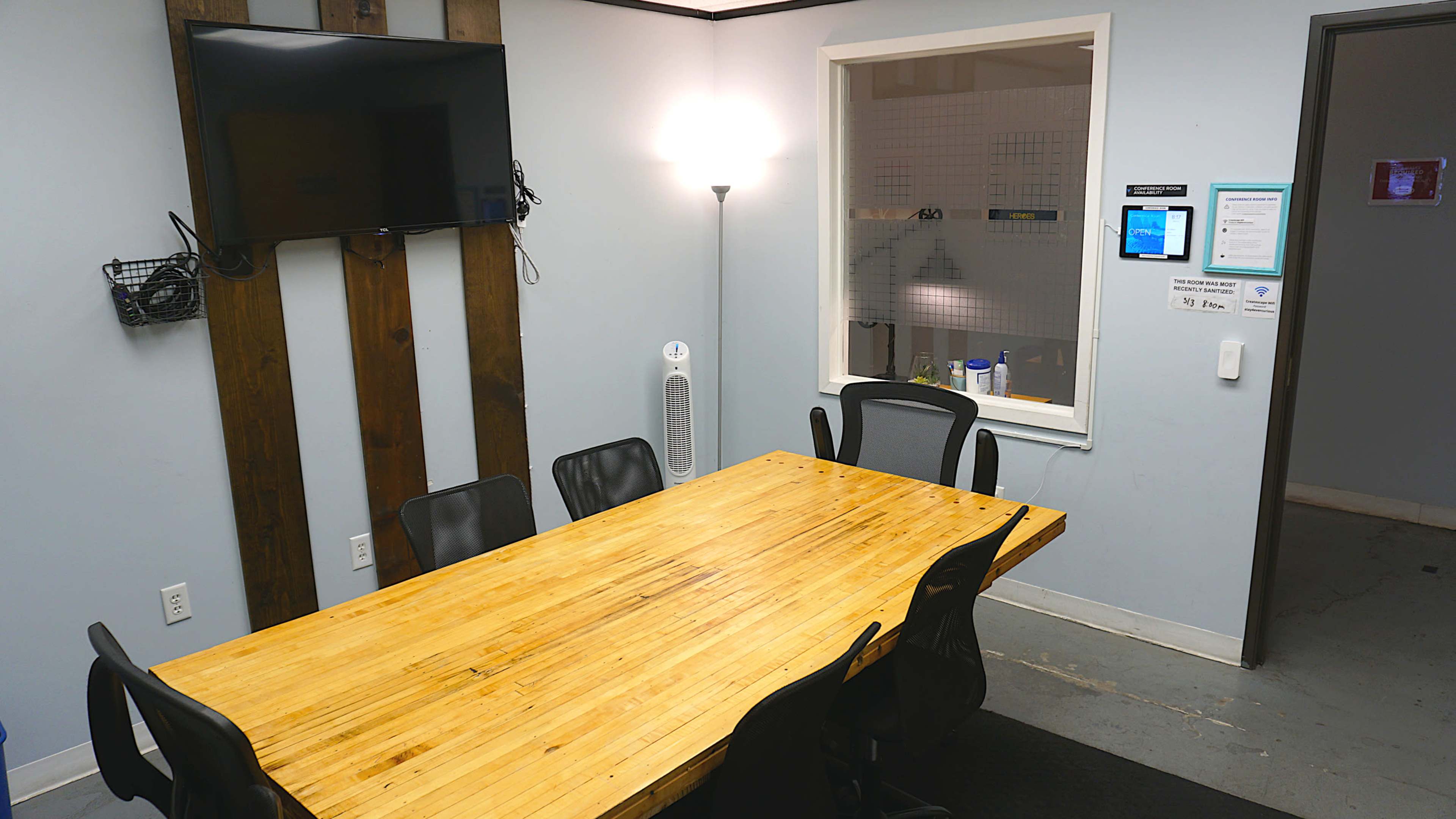 A meeting room with a wooden table surrounded by black chairs, a wall-mounted TV, and a window leading to an adjacent area.