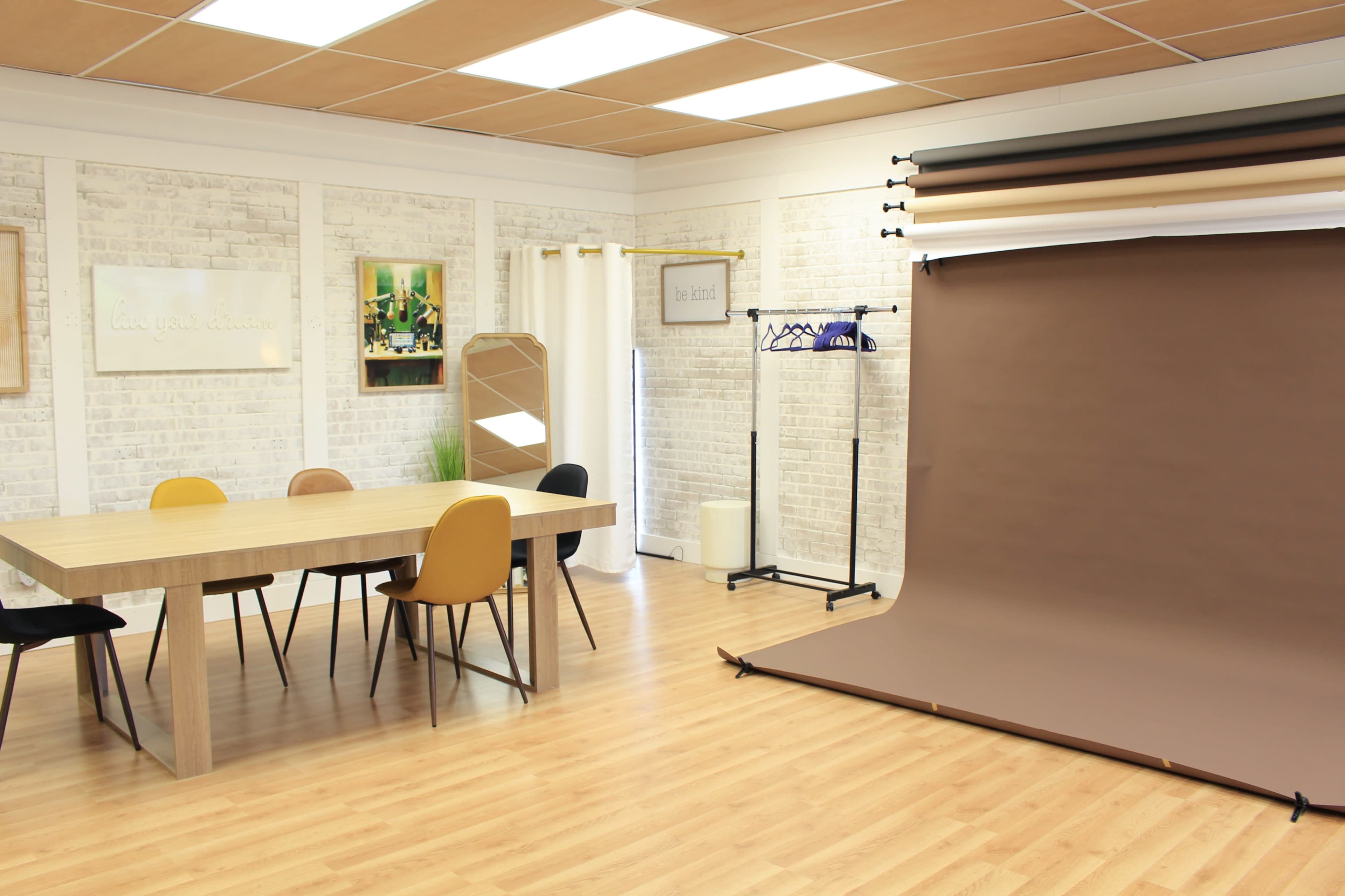 A bright, minimalist studio space with a wooden table, mixed-color chairs, and a backdrop stand featuring various fabric rolls.
