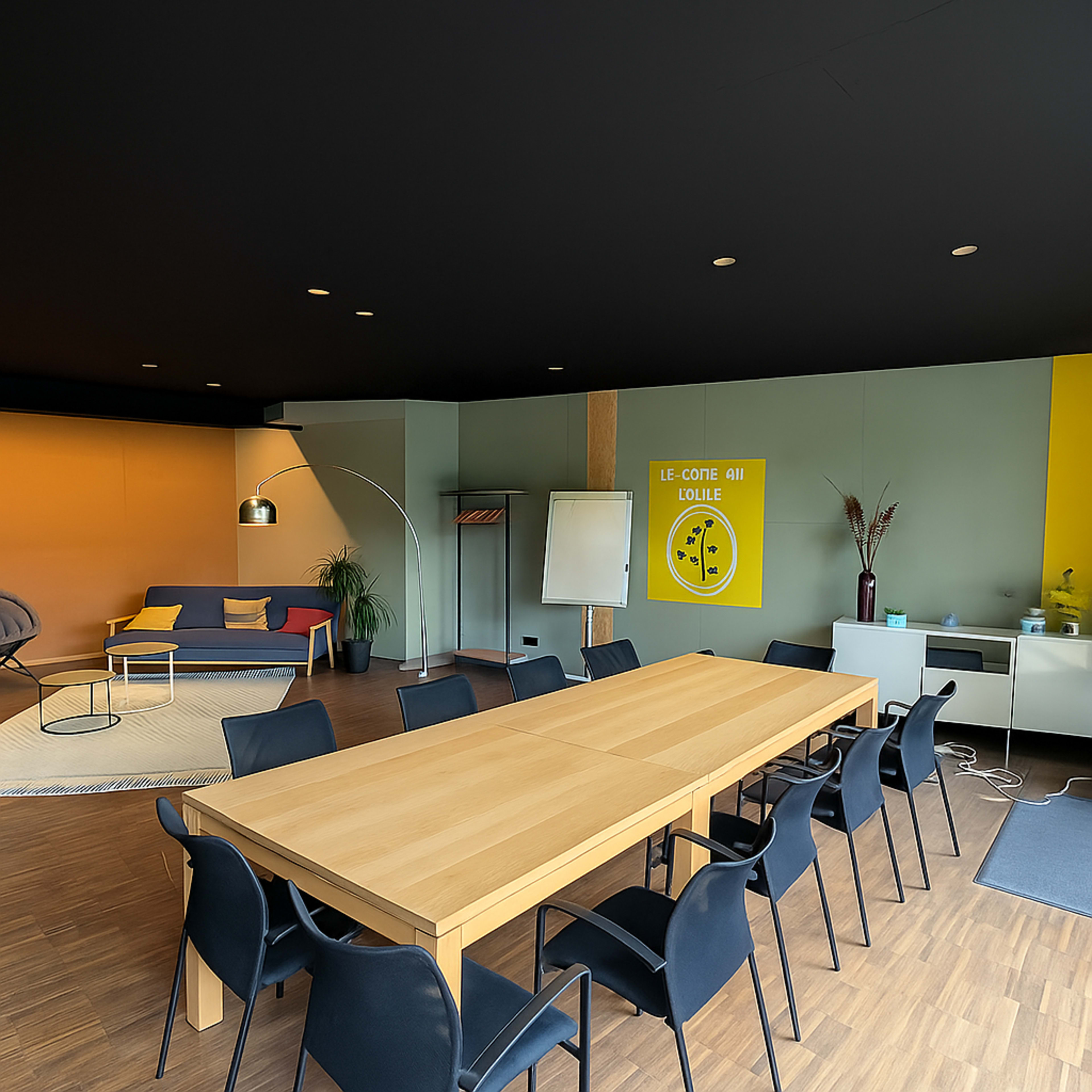 A spacious meeting room features a long wooden table surrounded by black chairs, with a colorful wall and a sofa in the background.