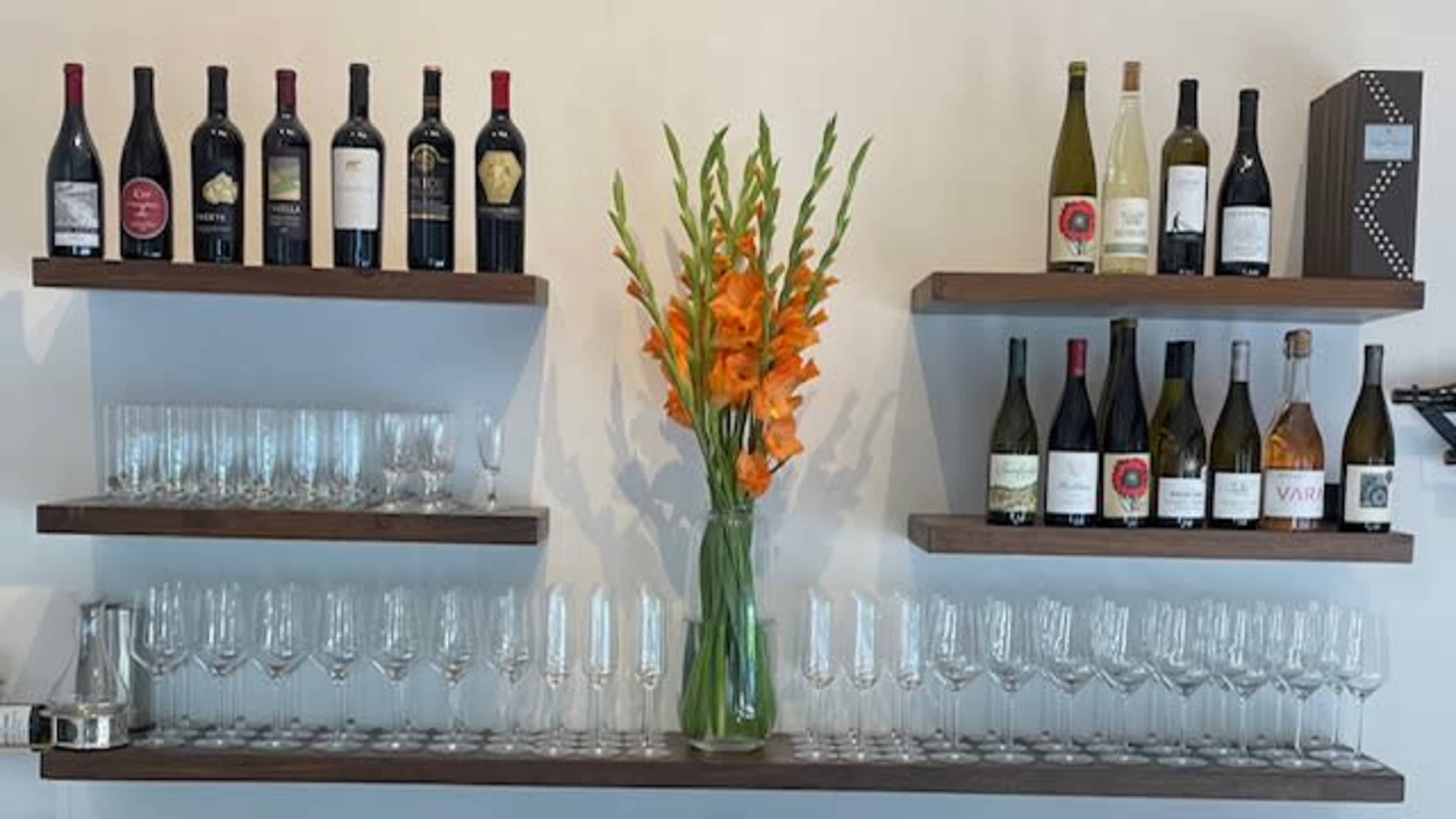 The image shows a wall-mounted display with two shelves containing various wine bottles and a central vase of orange flowers, complemented by rows of glasses stacked below.