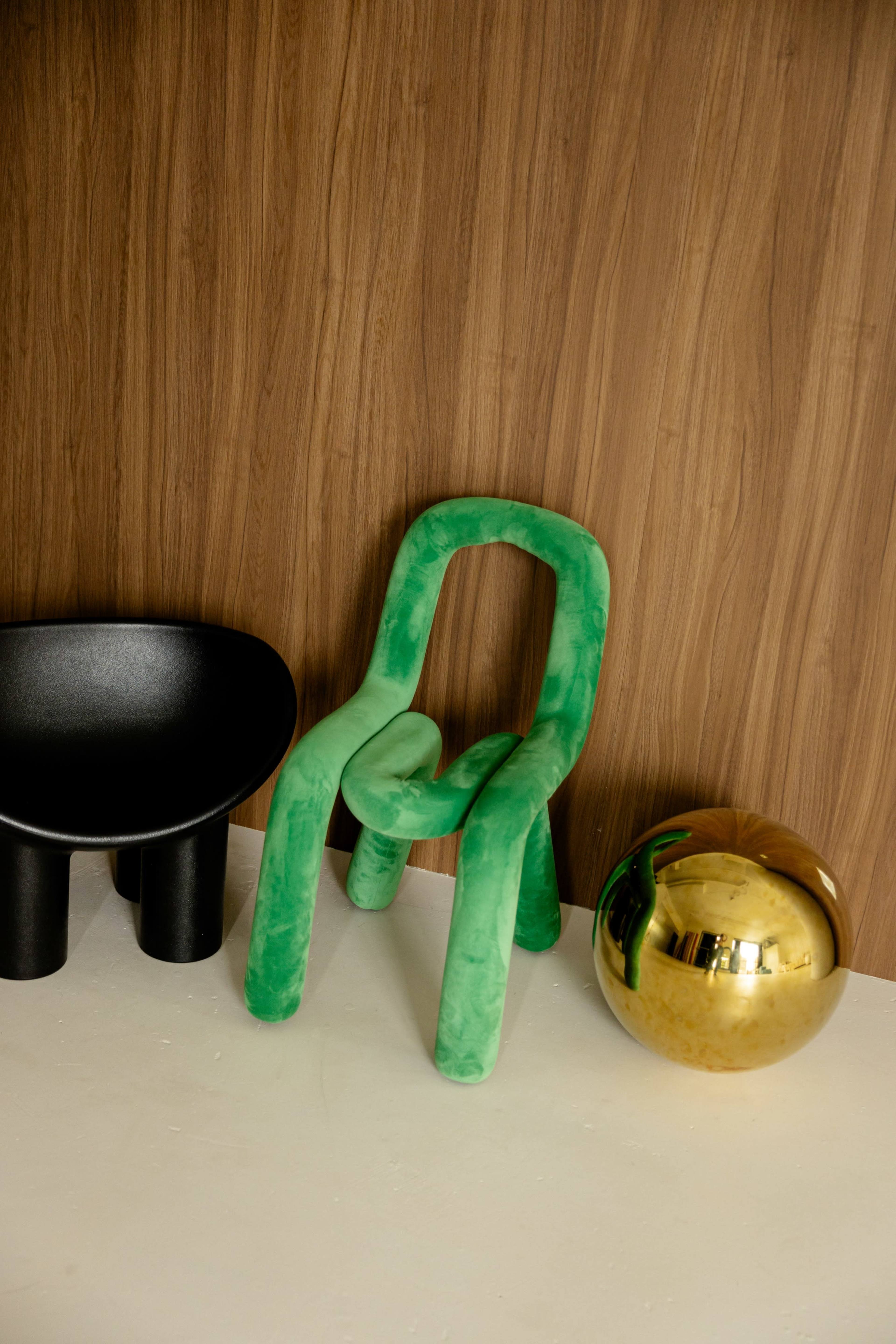 A green, sculptural chair is positioned next to a black bowl-shaped chair and a shiny gold spherical object against a wooden wall.