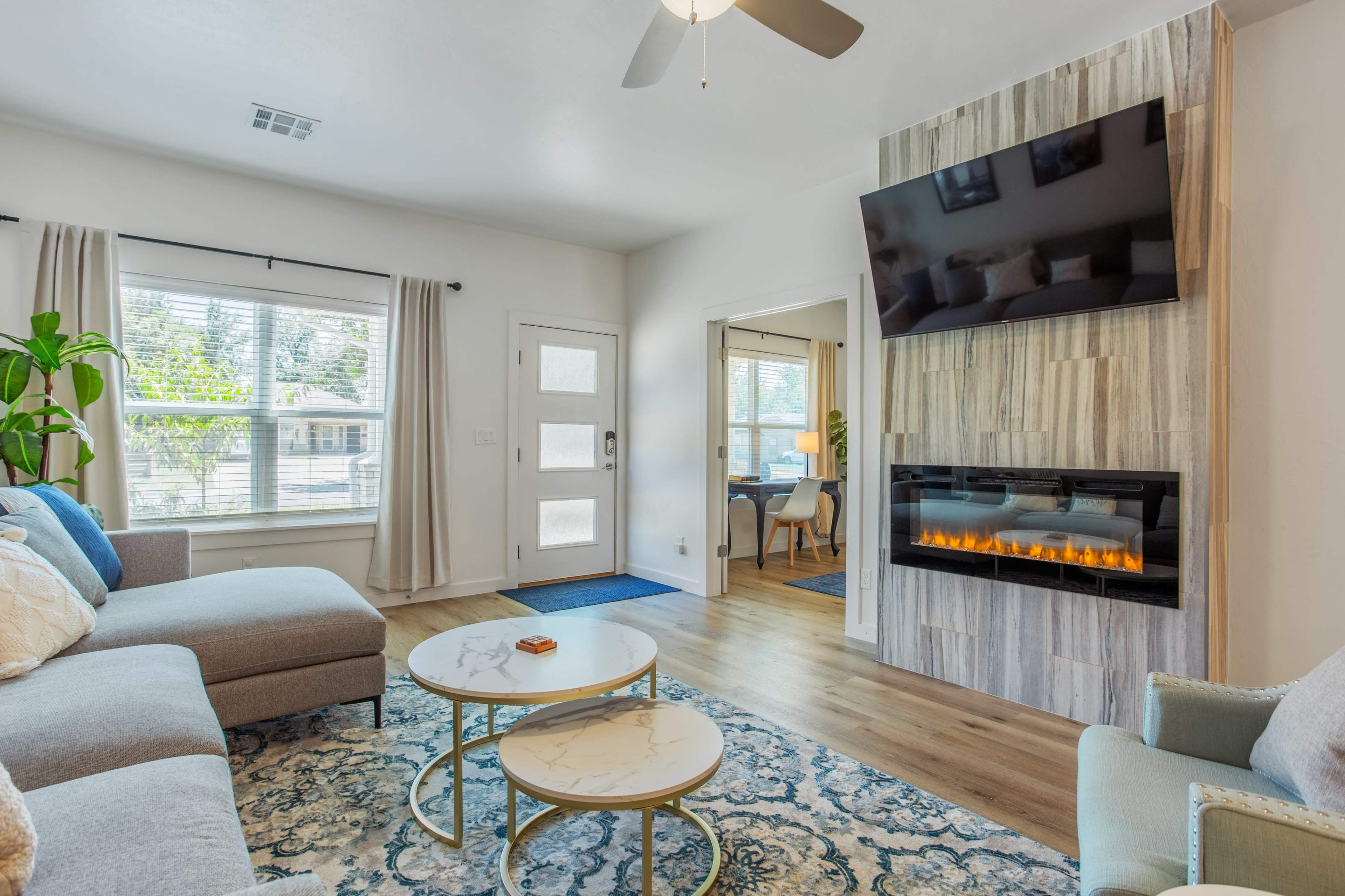 The living room features a sectional sofa, a wall-mounted television above a modern electric fireplace, and a pair of round coffee tables on a patterned rug.