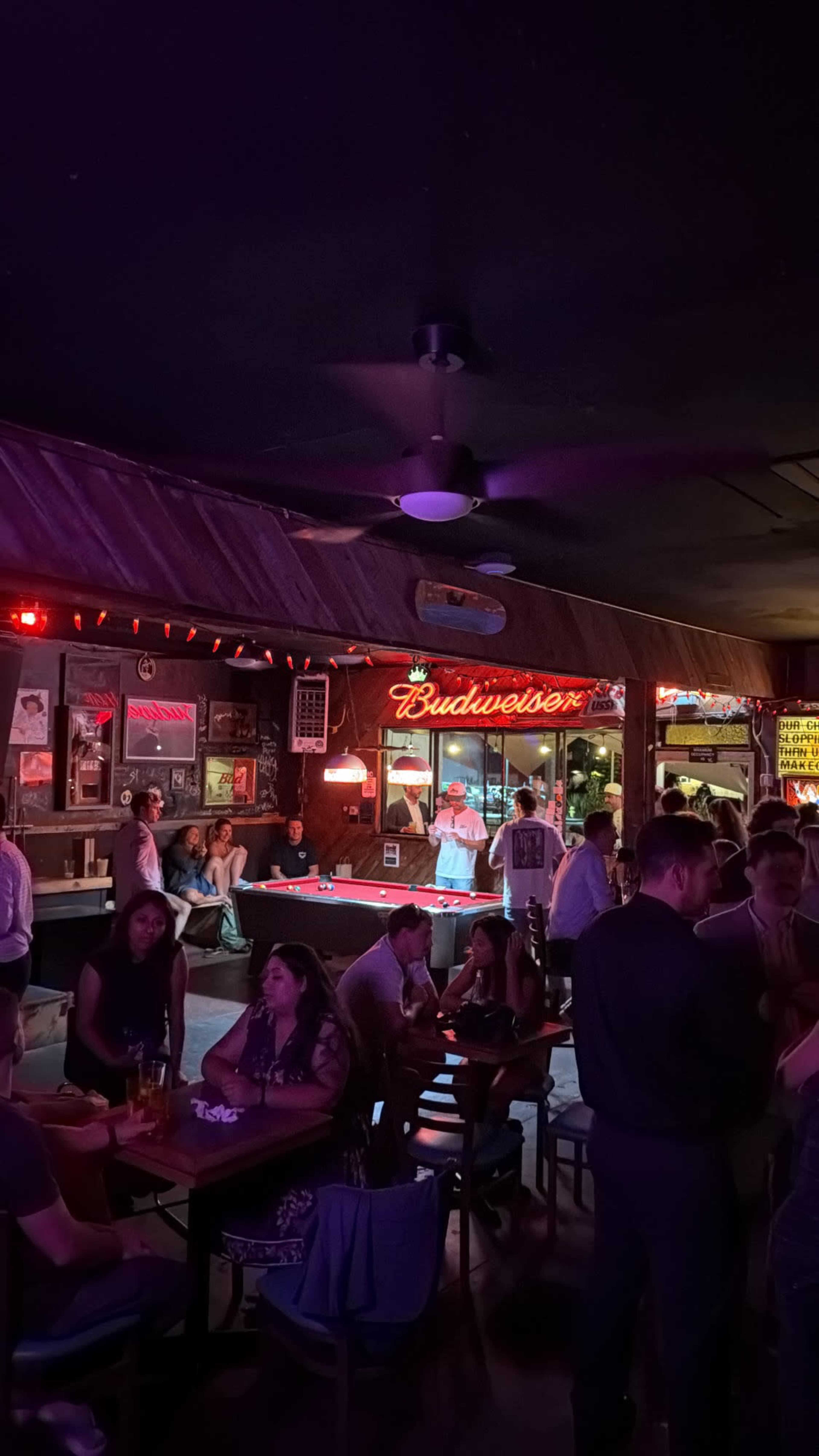 The interior of a dimly lit bar shows patrons gathered at tables, with a neon Budweiser sign illuminating the space.