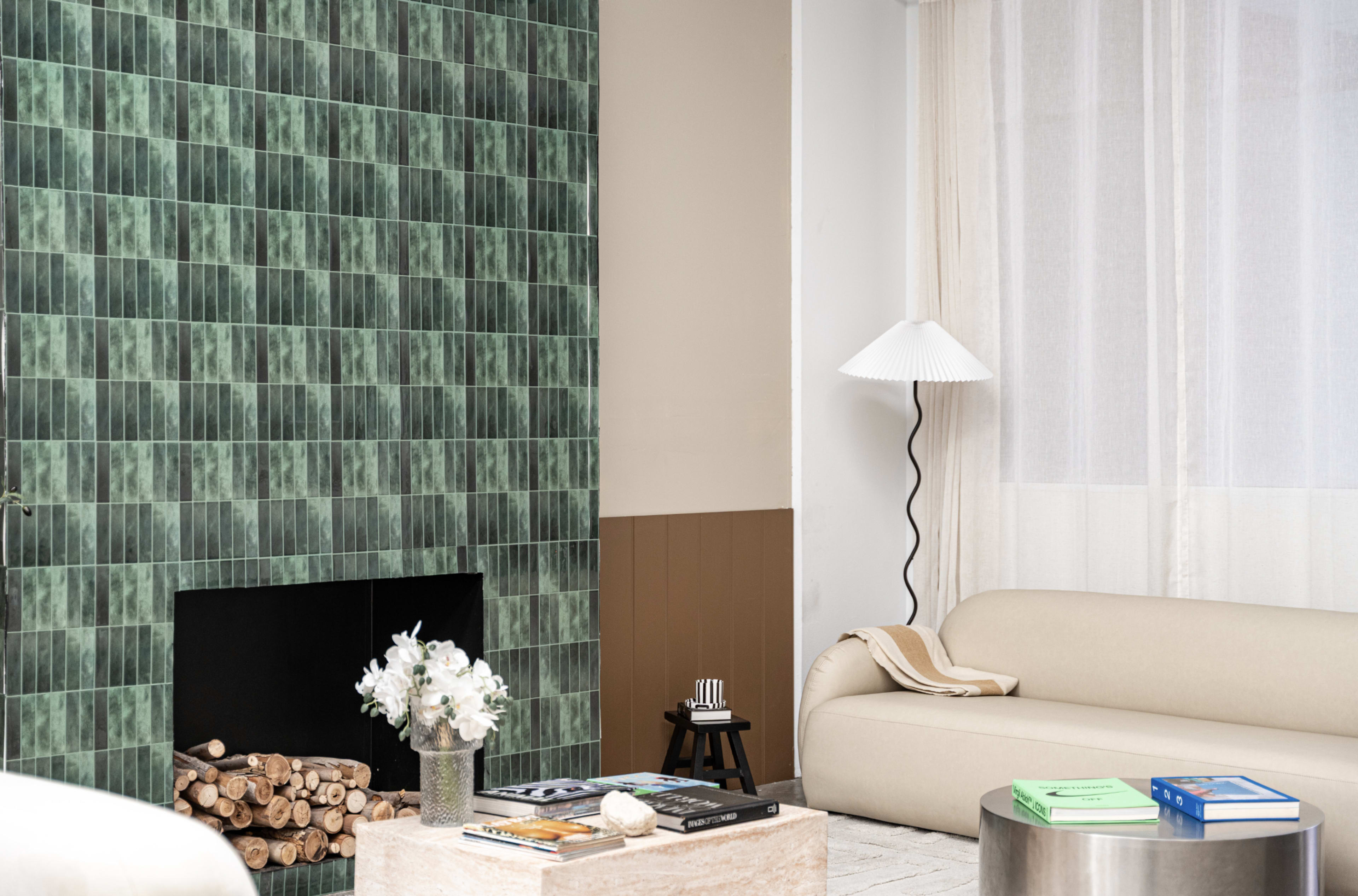 The image shows a modern living room featuring a green tiled wall with a fireplace, a beige sofa, a light-colored coffee table, and a floor lamp, illuminated by natural light through sheer curtains.
