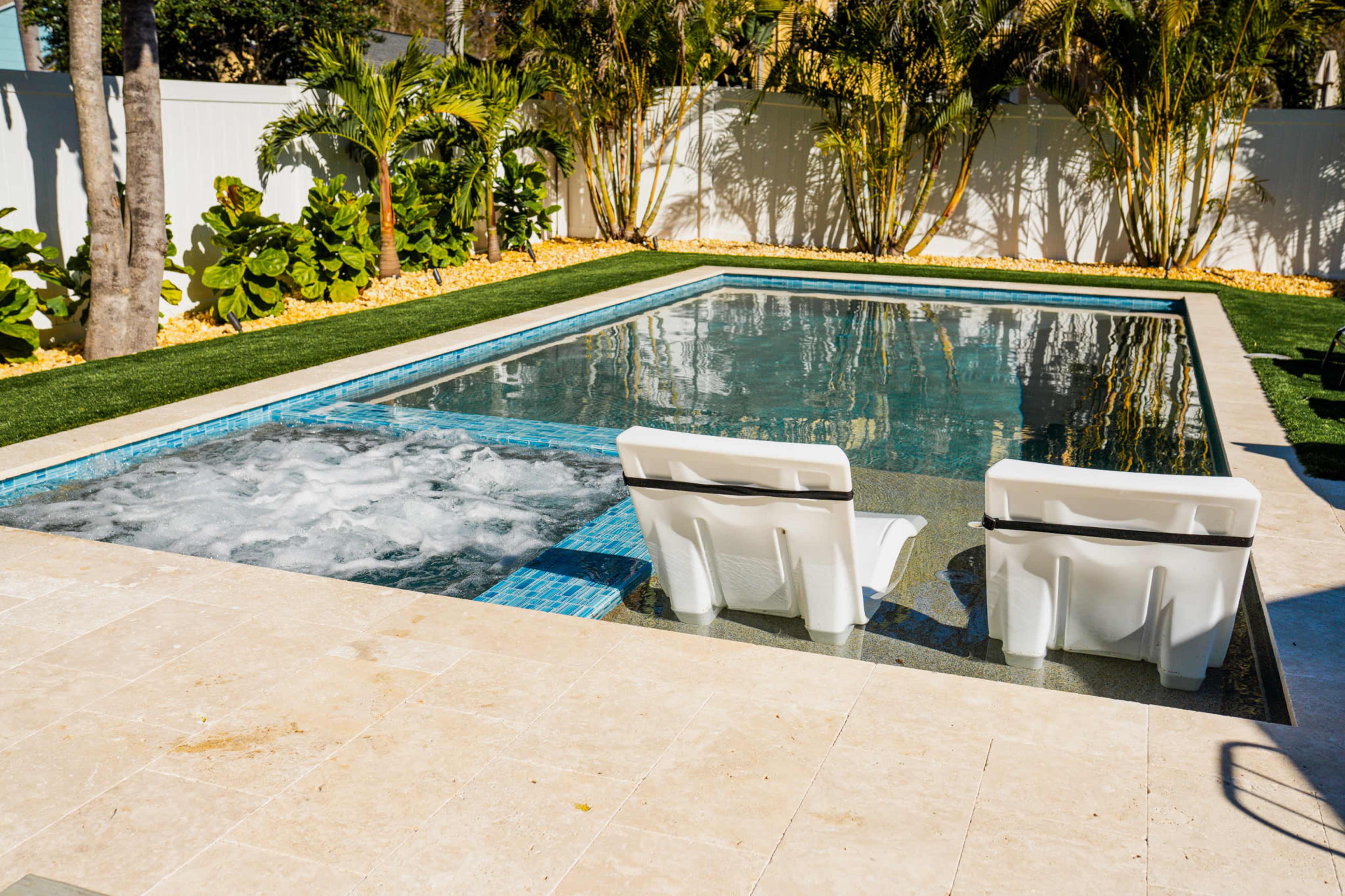 A rectangular swimming pool features a jacuzzi area and two white lounge chairs positioned partially in the water, surrounded by greenery and a paved deck.
