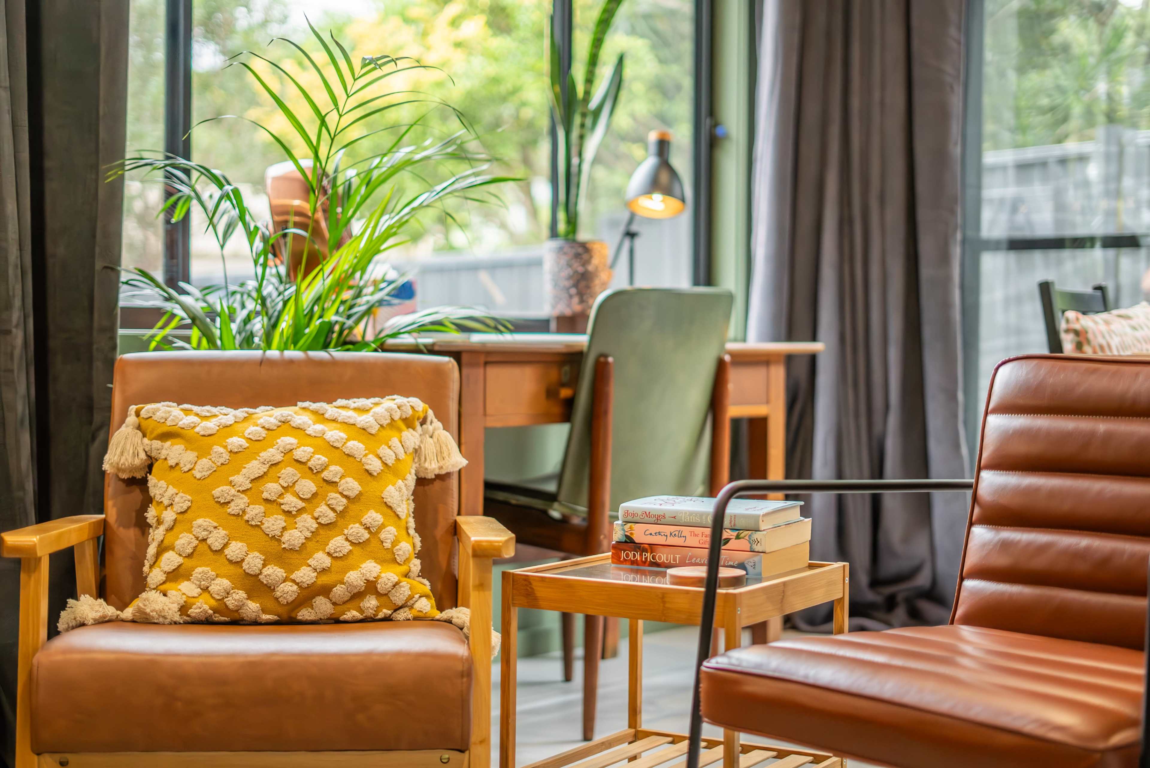 The image shows a cozy living space with two armchairs, a small wooden table with stacked books, and a desk in the background, surrounded by large windows and indoor plants.