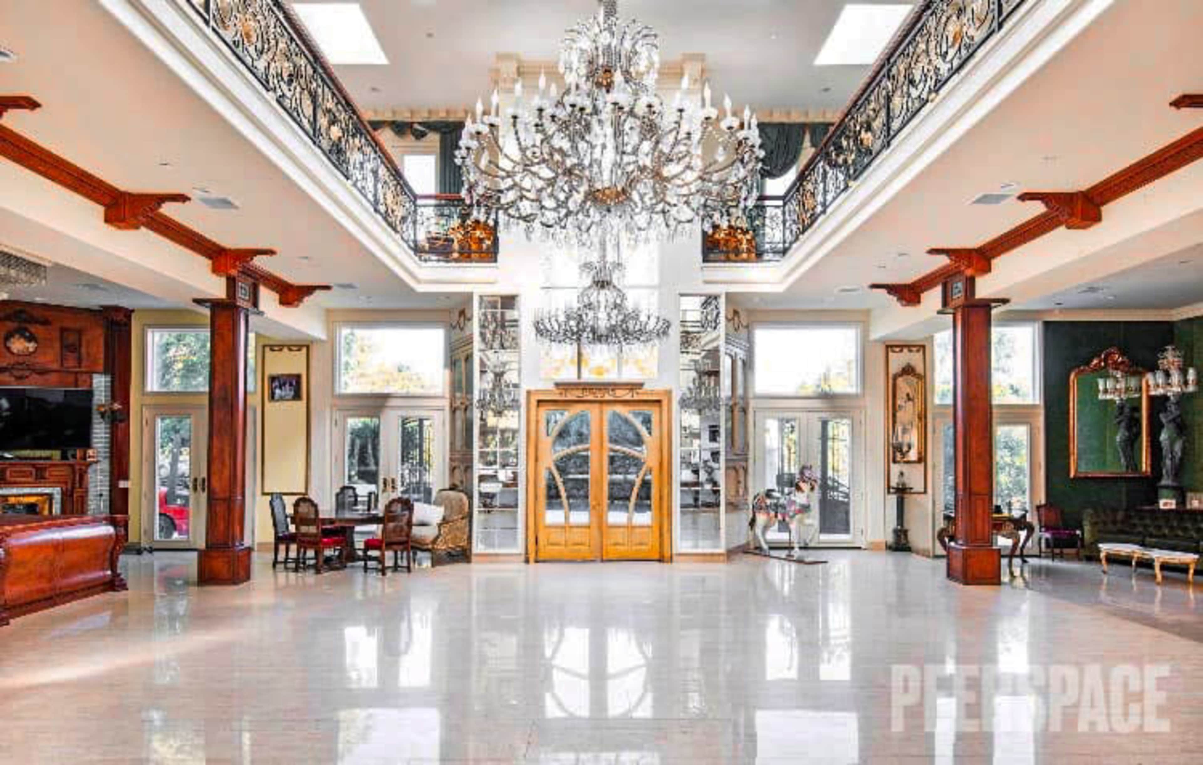 An expansive foyer with a high ceiling, featuring a grand chandelier, elegant wooden doors, and a polished marble floor.