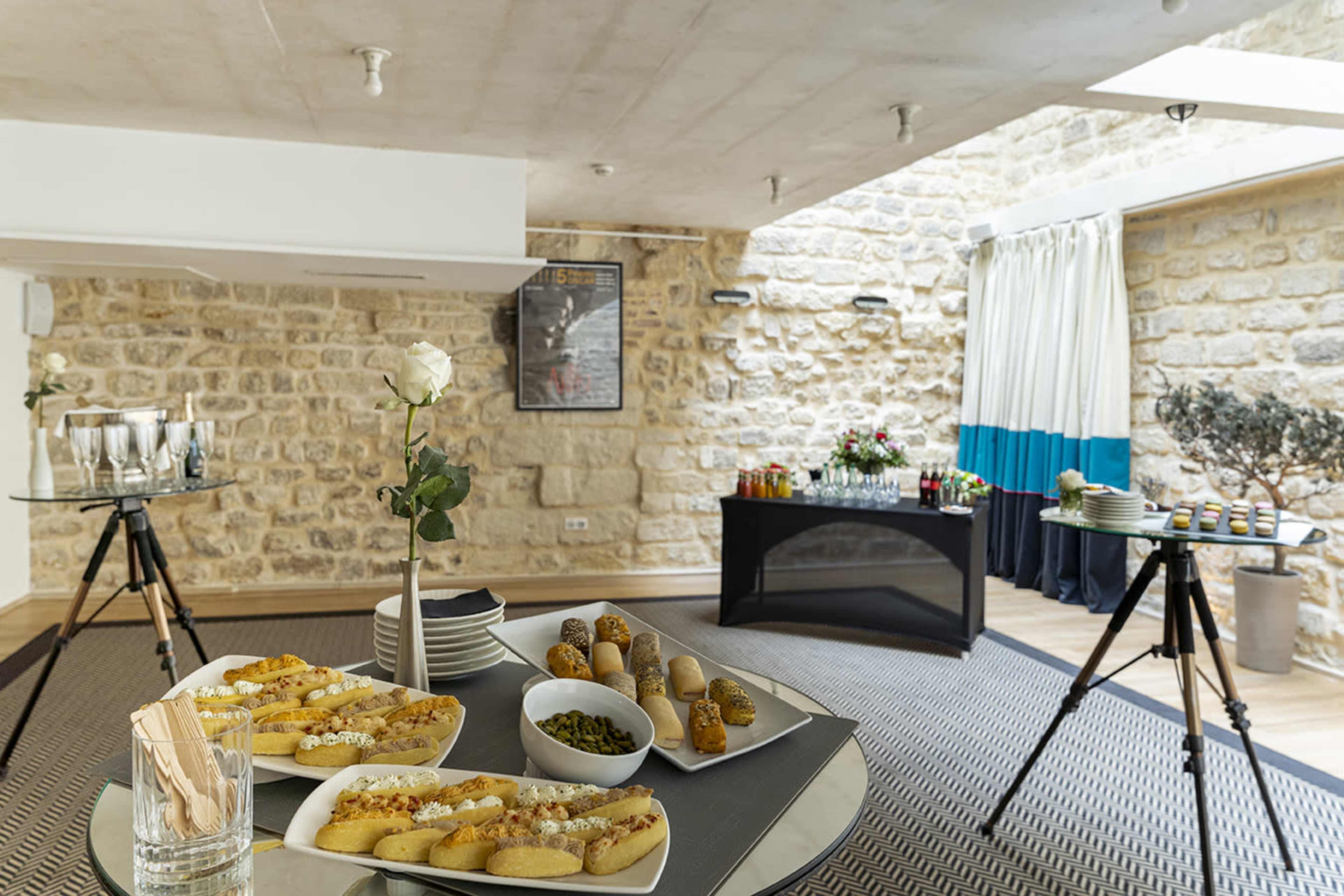 A neatly arranged event space features tables with appetizers and drinks against a backdrop of stone walls and large windows.