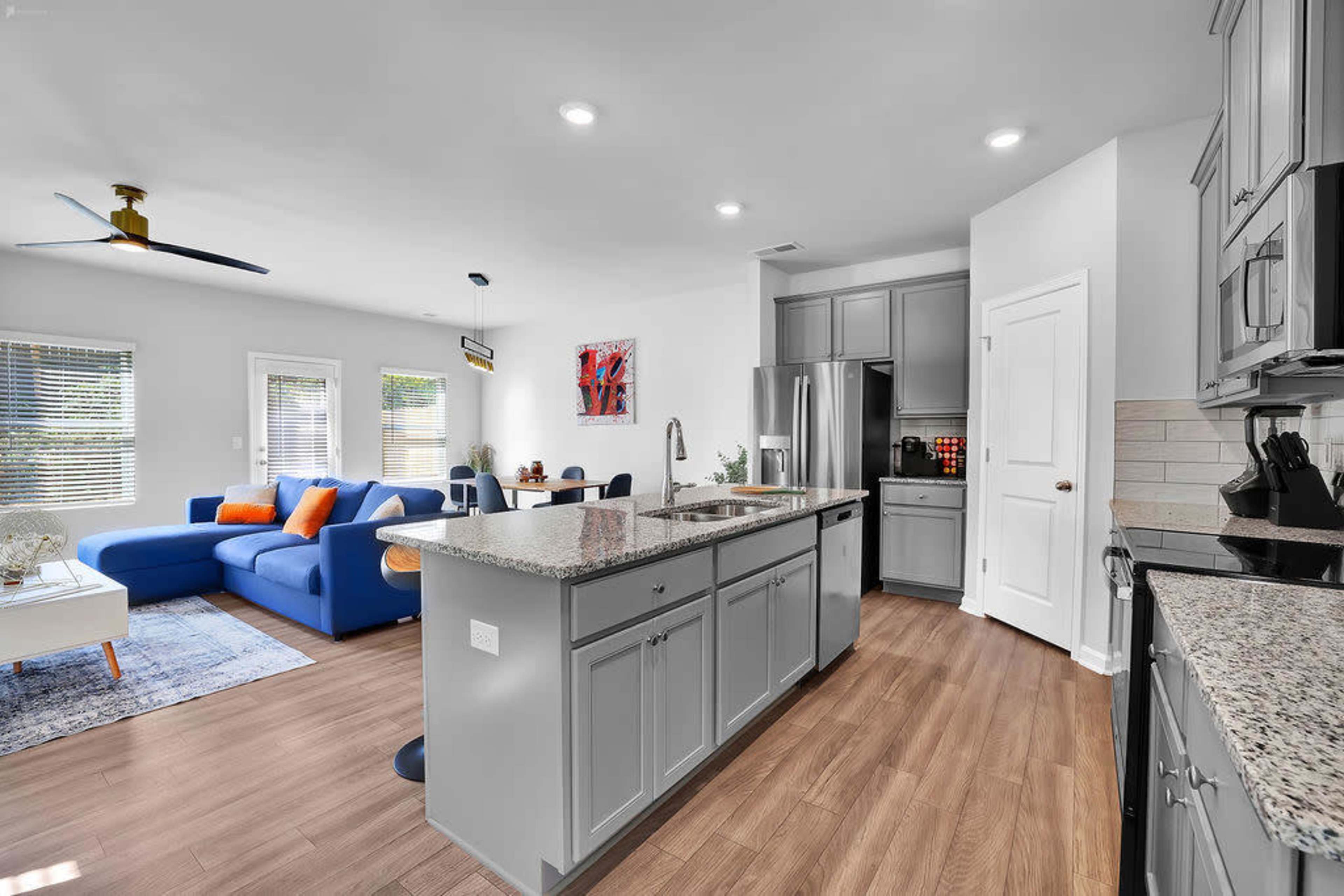 A modern kitchen with gray cabinets, a granite countertop, and an open living area featuring a blue sofa and dining space.