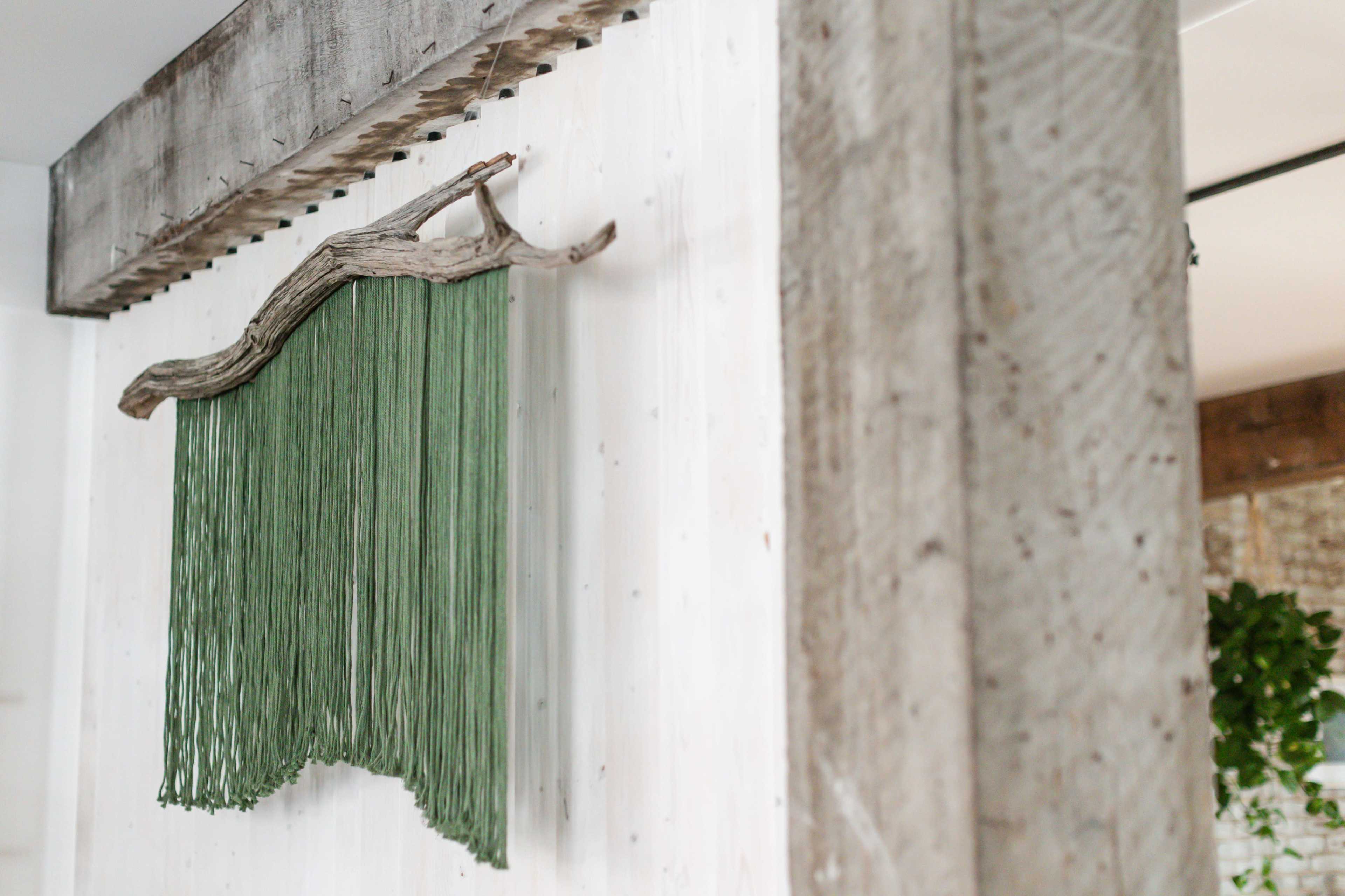 A piece of green wall art hangs from a wooden branch against a white wall.