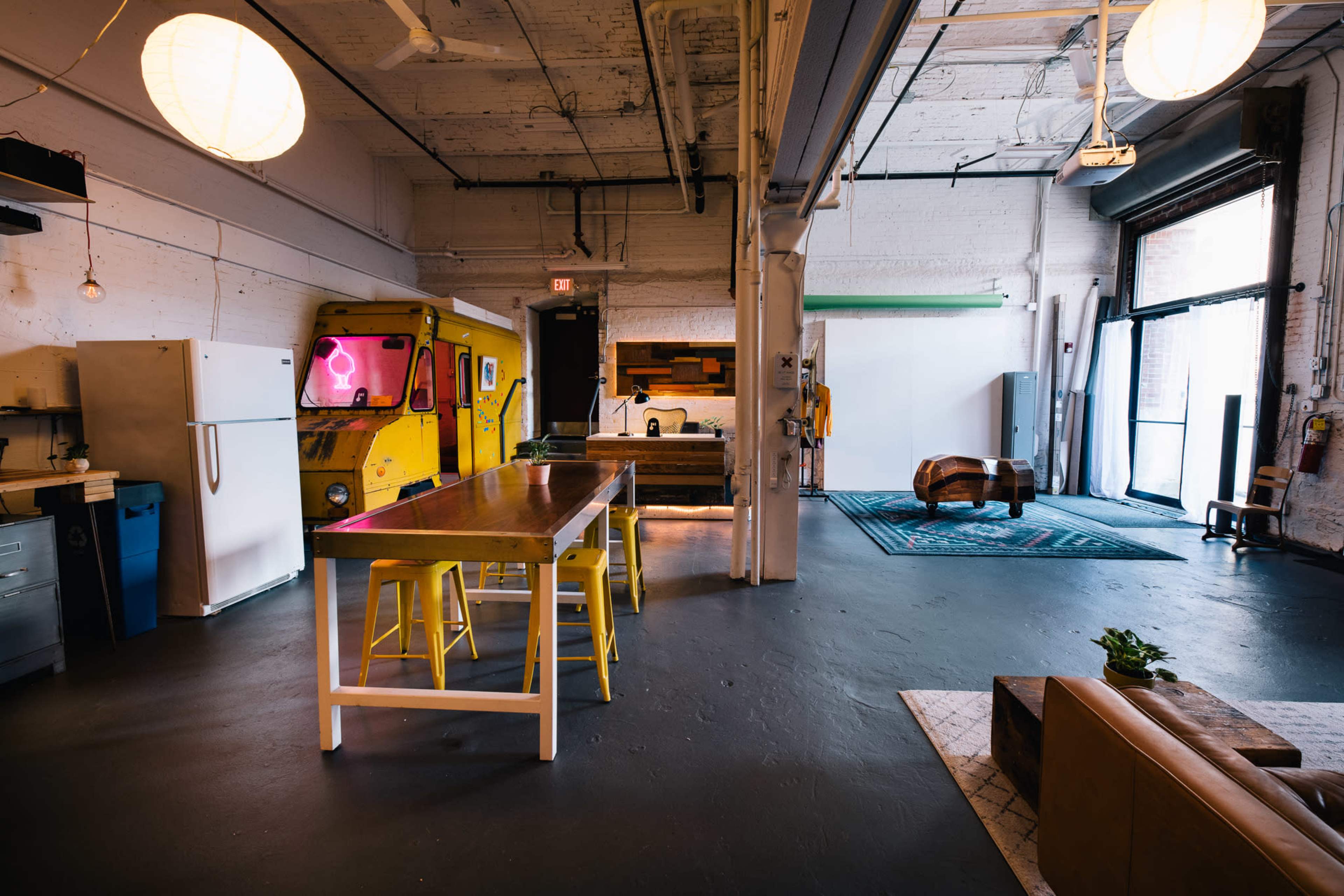A spacious, well-lit industrial-style room features a yellow food truck, a long dining table with yellow stools, various kitchen appliances, and a relaxed seating area.