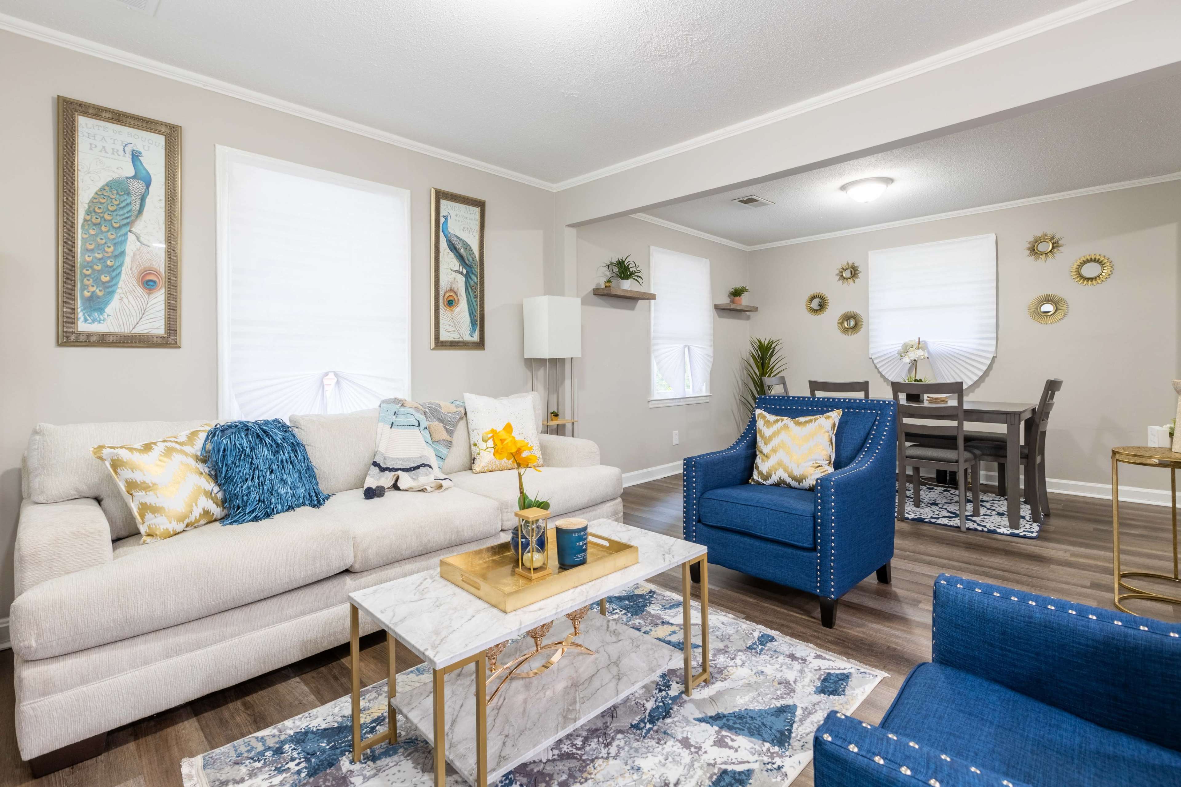A living room features a light-colored sofa, a blue armchair, a marble coffee table, and a dining area with a table and chairs, adorned with decorative wall art and plants.