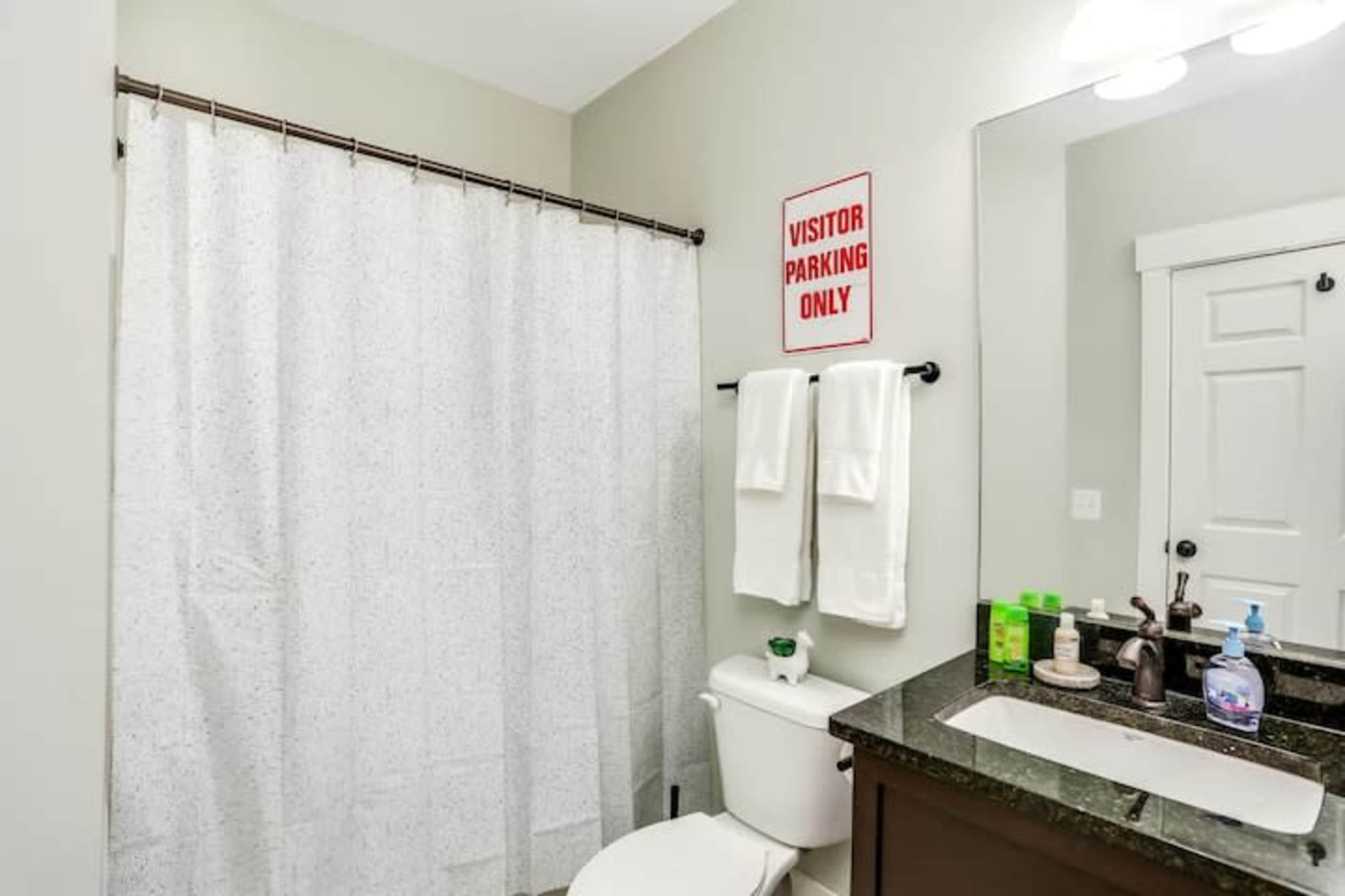 A bathroom features a white shower curtain, a toilet, a sink with a dark countertop, and a red "Visitor Parking Only" sign on the wall.
