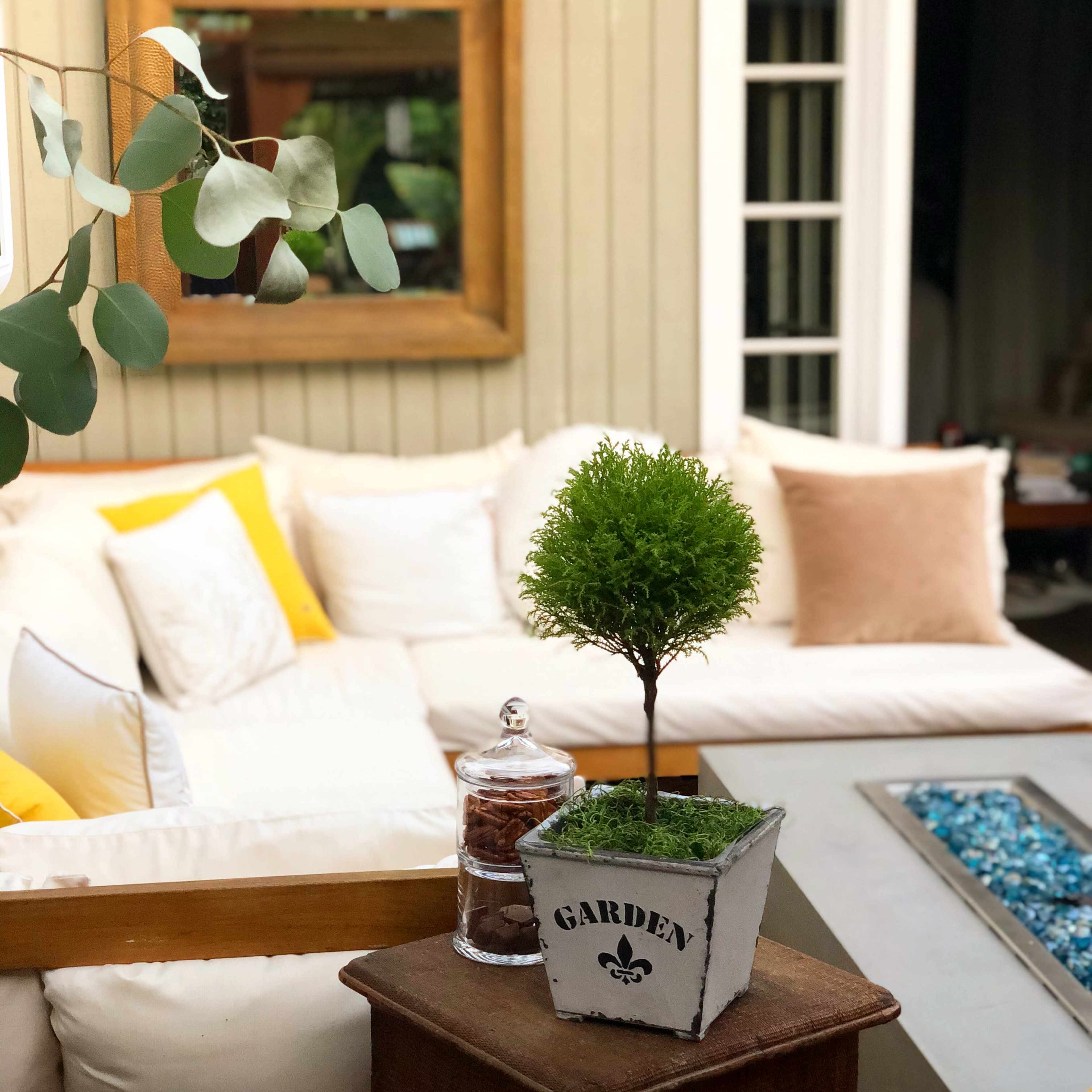 A cozy outdoor seating area featuring a white couch with colorful cushions, a small potted tree, and a glass jar beside a decorative planter on a wooden table.