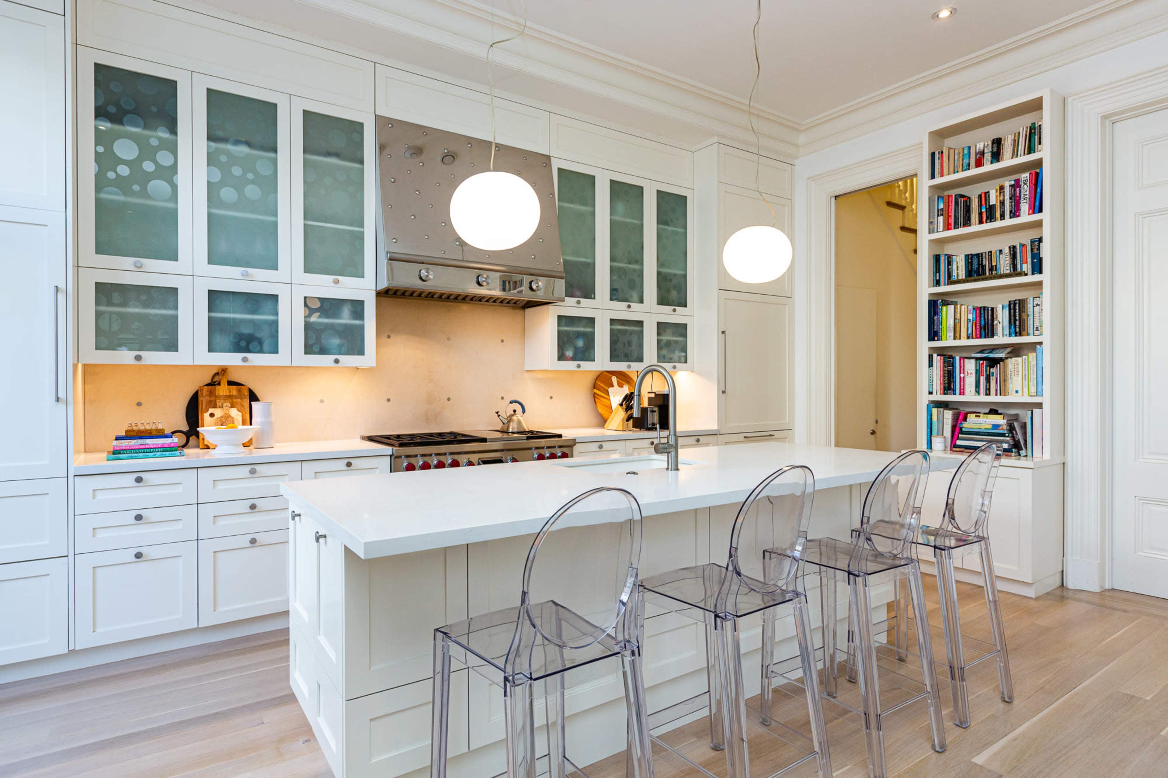 The image depicts a modern kitchen with white cabinetry, a central island with transparent stools, and a stainless steel range hood.