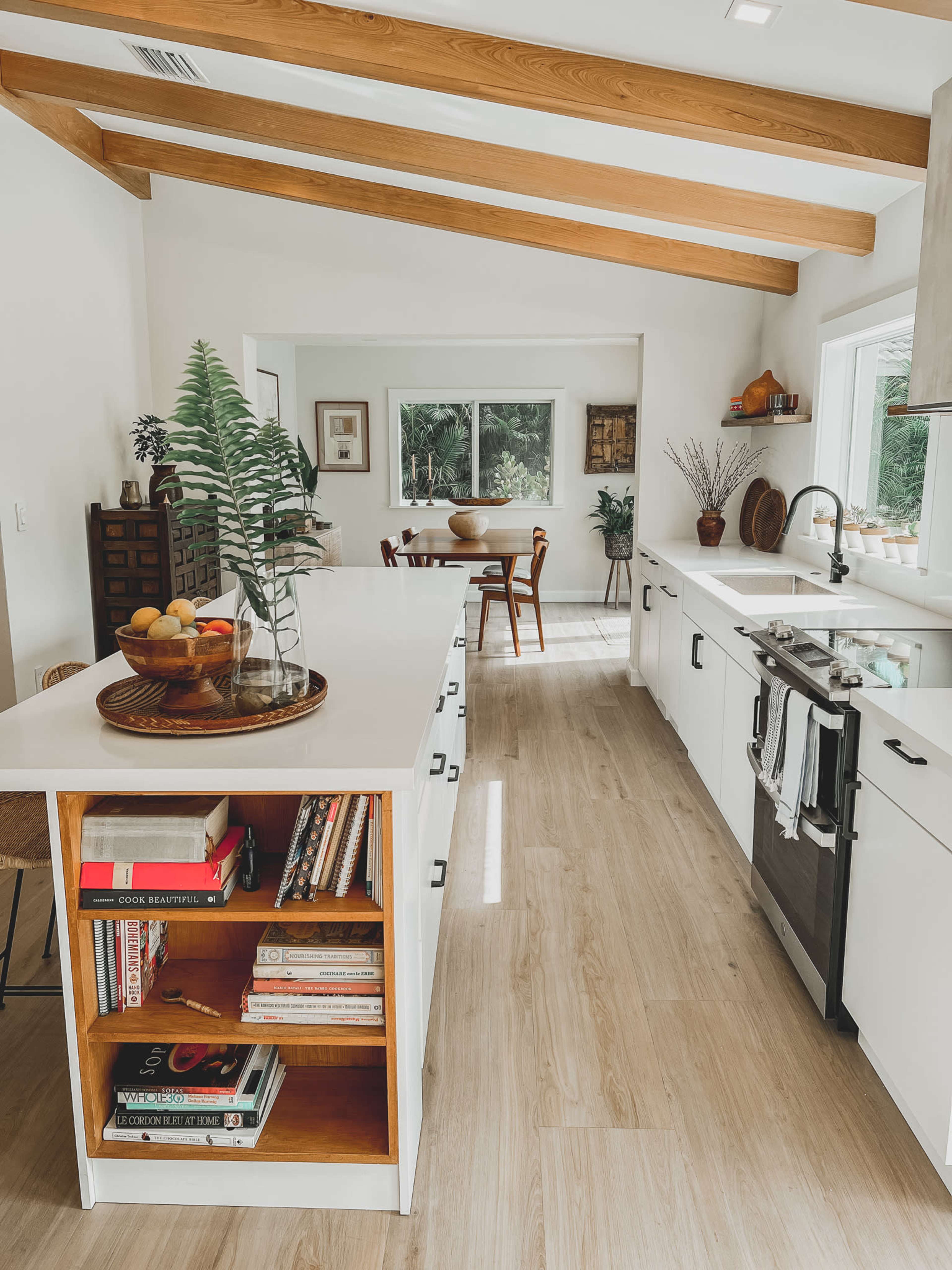 A modern kitchen features a central island with shelves of books, white cabinets, and wooden beams overhead, leading into a dining area with chairs and a window view.