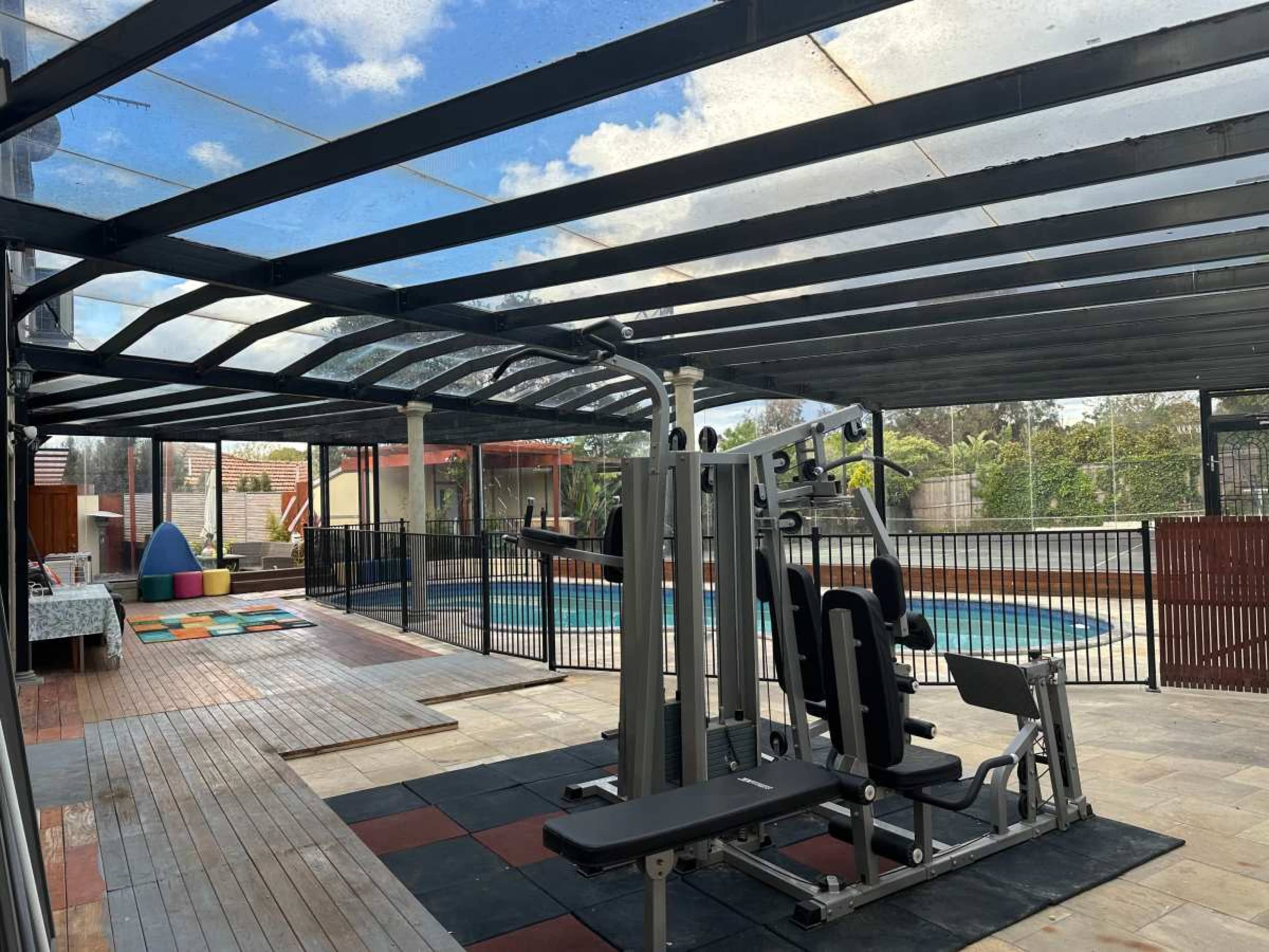 The image shows a covered outdoor area featuring a gym equipment setup adjacent to a swimming pool.