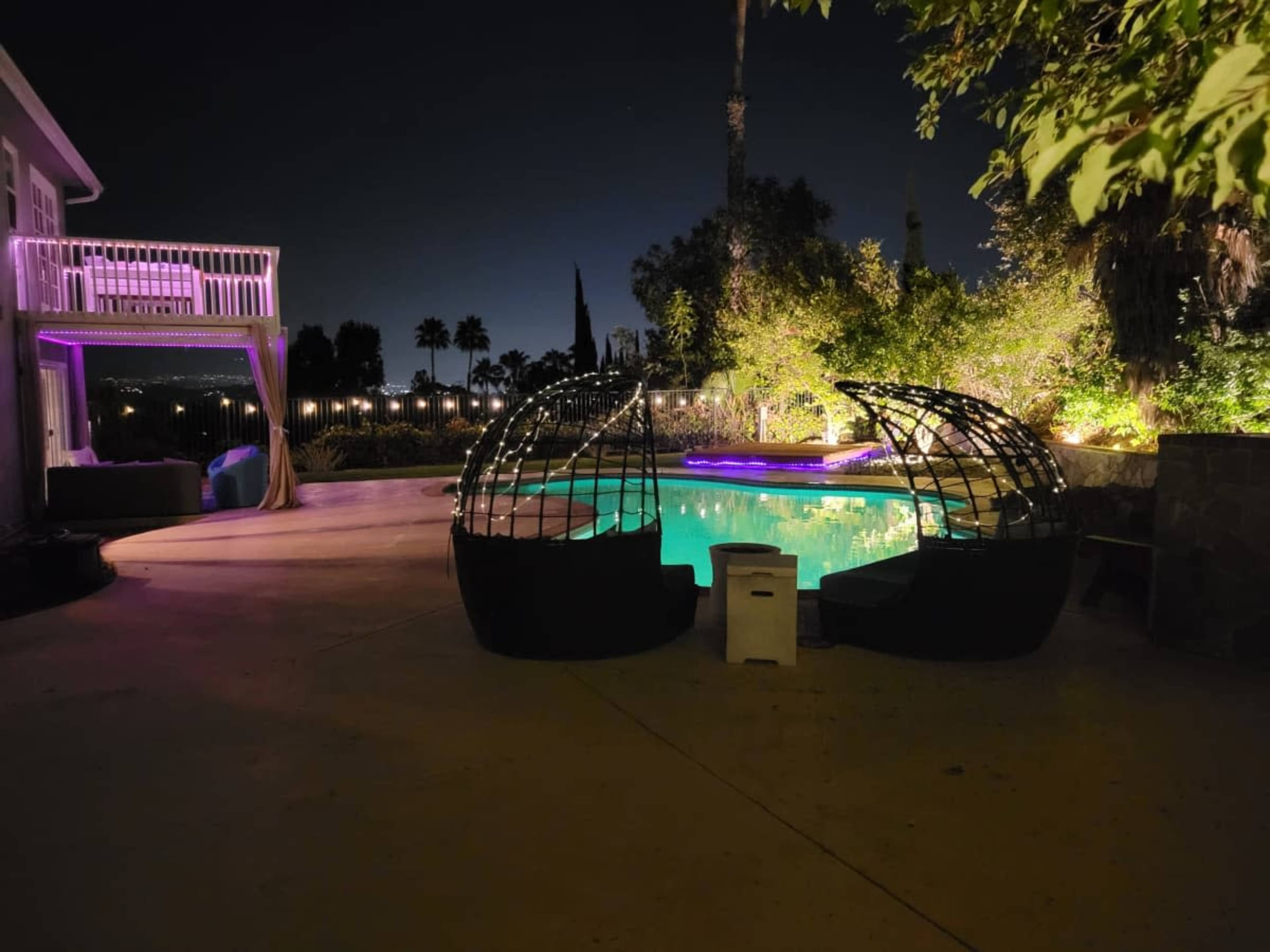 The image shows a nighttime view of a outdoor pool area featuring two curved lounge structures near the water, illuminated by soft purple lights, with a house and palm trees in the background.