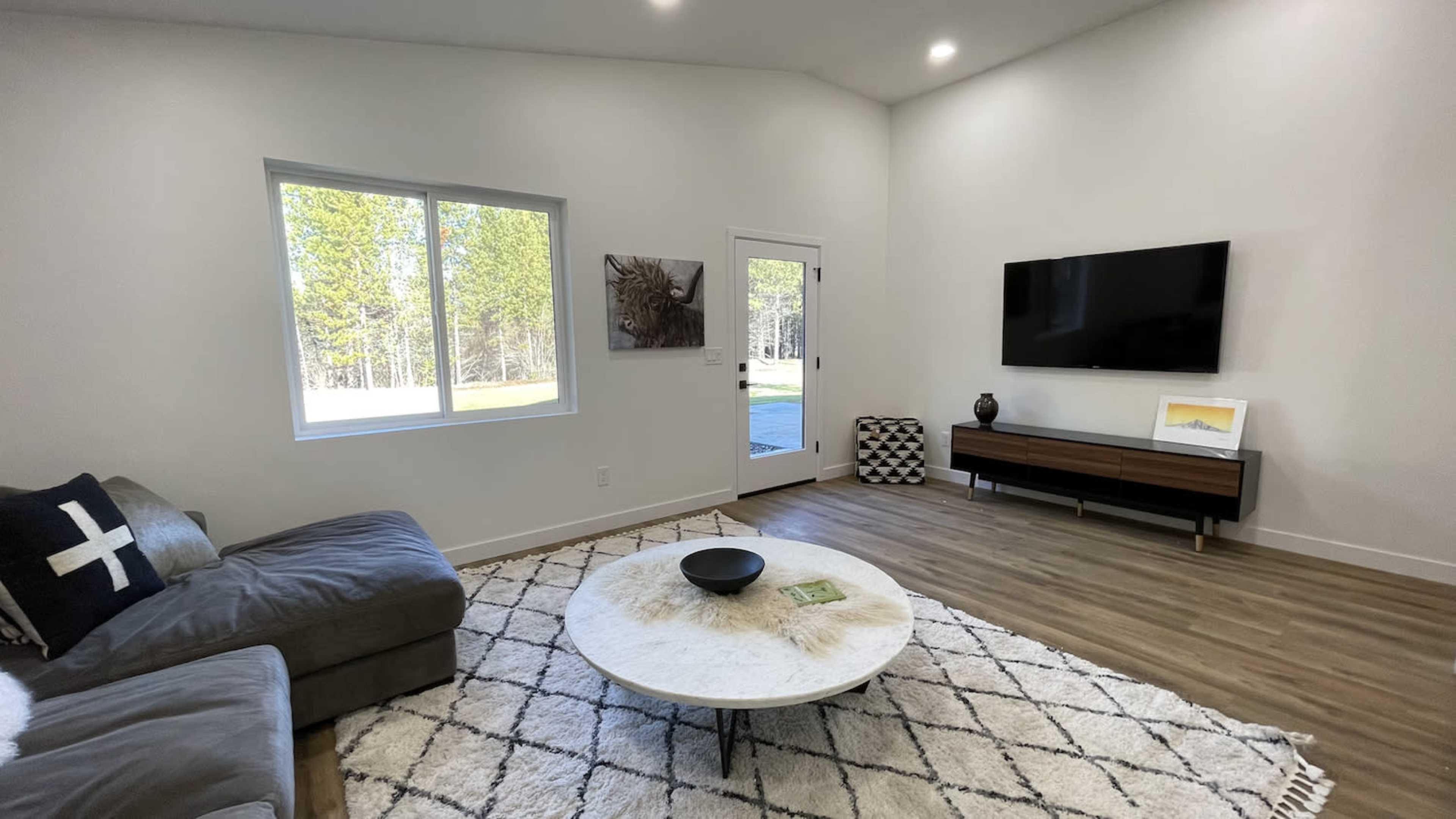 A modern living room features a gray sectional sofa, a round coffee table on a textured rug, a wall-mounted TV, and large windows overlooking a wooded area.